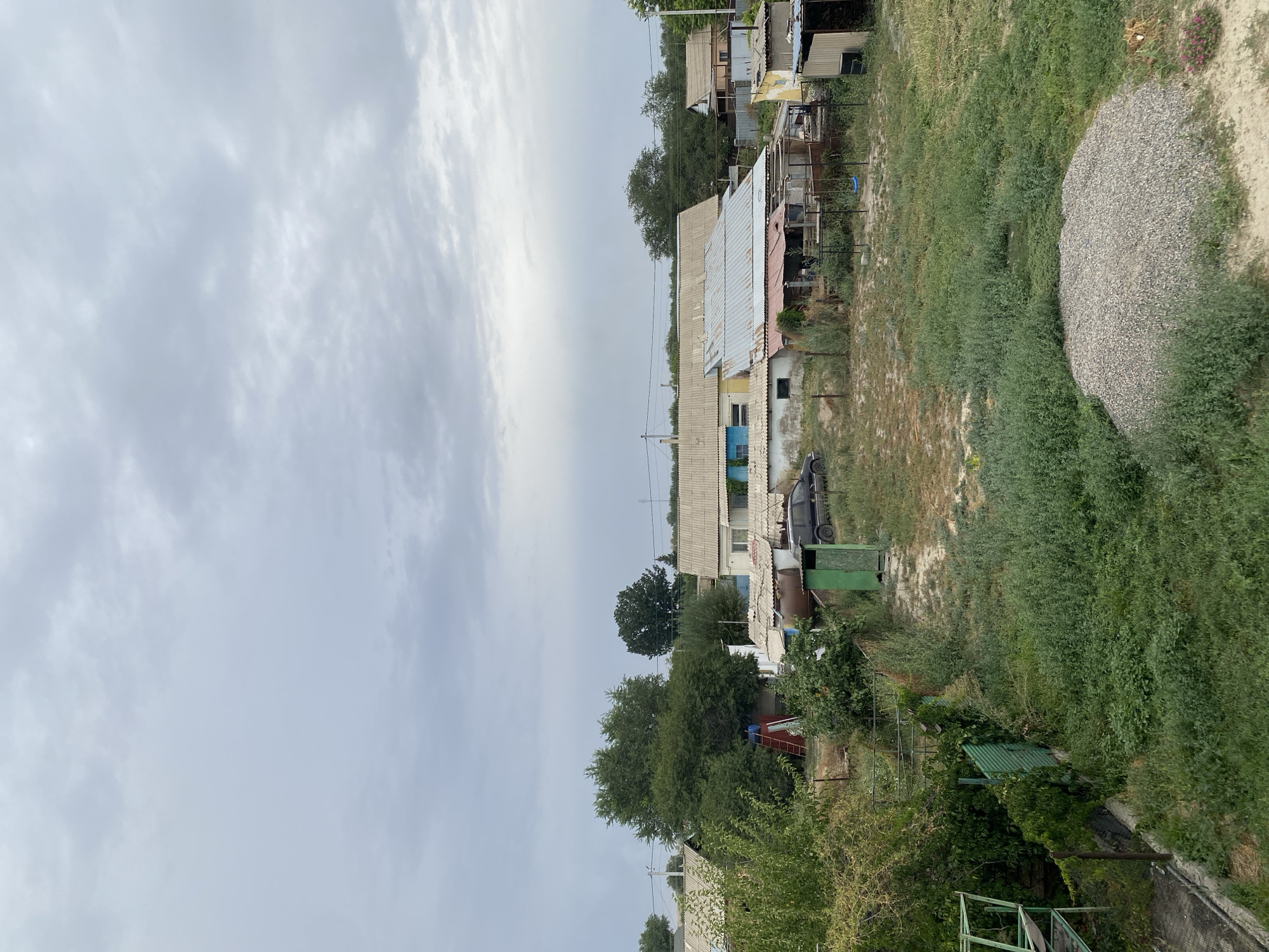 The image depicts a cloudy sky over a residential area in Qaraoy, Kazakhstan. In the foreground, a pile of gravel sits amidst overgrown green vegetation. A green metal fence runs along the left side, partially covered in vines. Moving towards the midground, more green foliage and a grassy yard are visible. Several low-rise buildings with corrugated metal roofs are scattered throughout the scene. One building has a blue window. A dark-colored car is parked near one of the structures. A red ladder leans against a large tree. A small green outhouse-like structure stands in the yard. In the background, more trees and buildings are visible under the overcast sky.