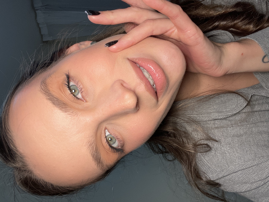 A woman's face, tilted, fills the frame. She gazes upward with light green eyes, accented by filled-in eyebrows and mascara. Her skin is smooth and seemingly flawless, with full, glossy lips. One hand is raised, fingers near her forehead. The nails are painted dark with a glitter effect. She wears a gray, textured shirt. Dark brown, wavy hair frames her face. A crescent moon tattoo is visible on her wrist. The background is a soft, muted gray-blue.