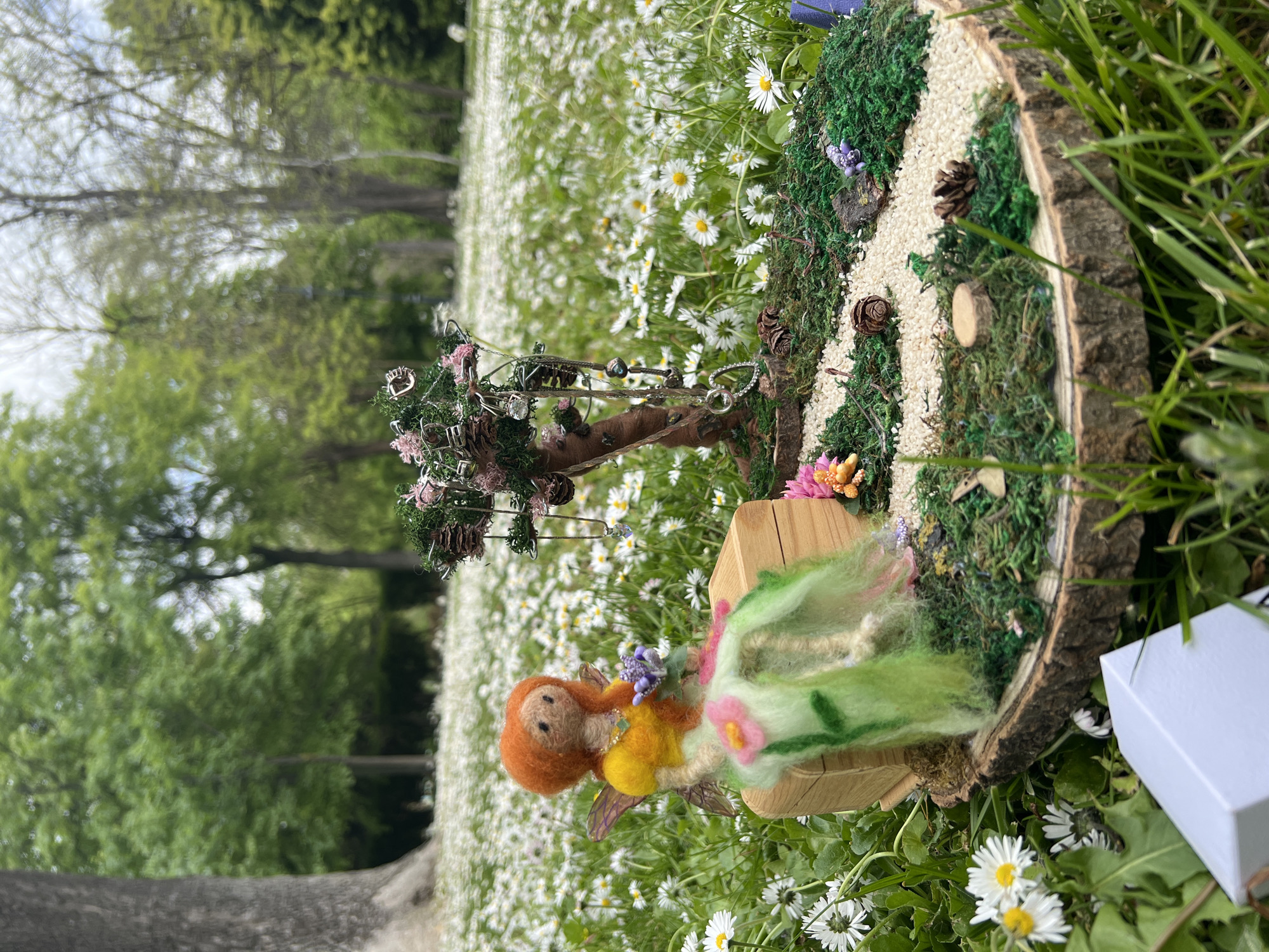 A close-up shot shows a fairy garden scene placed on a grassy field of daisies. A felted doll with red hair, a yellow bodice, and a green skirt sits on a small wooden stool. A miniature tree, adorned with jewelry and pink flowers, stands behind the doll. A small, circular stage made from a tree slice sits nearby, decorated with moss, pinecones, and a path of white pebbles. The background is a blur of trees and the daisy field. The location is Burgas, Bulgaria.