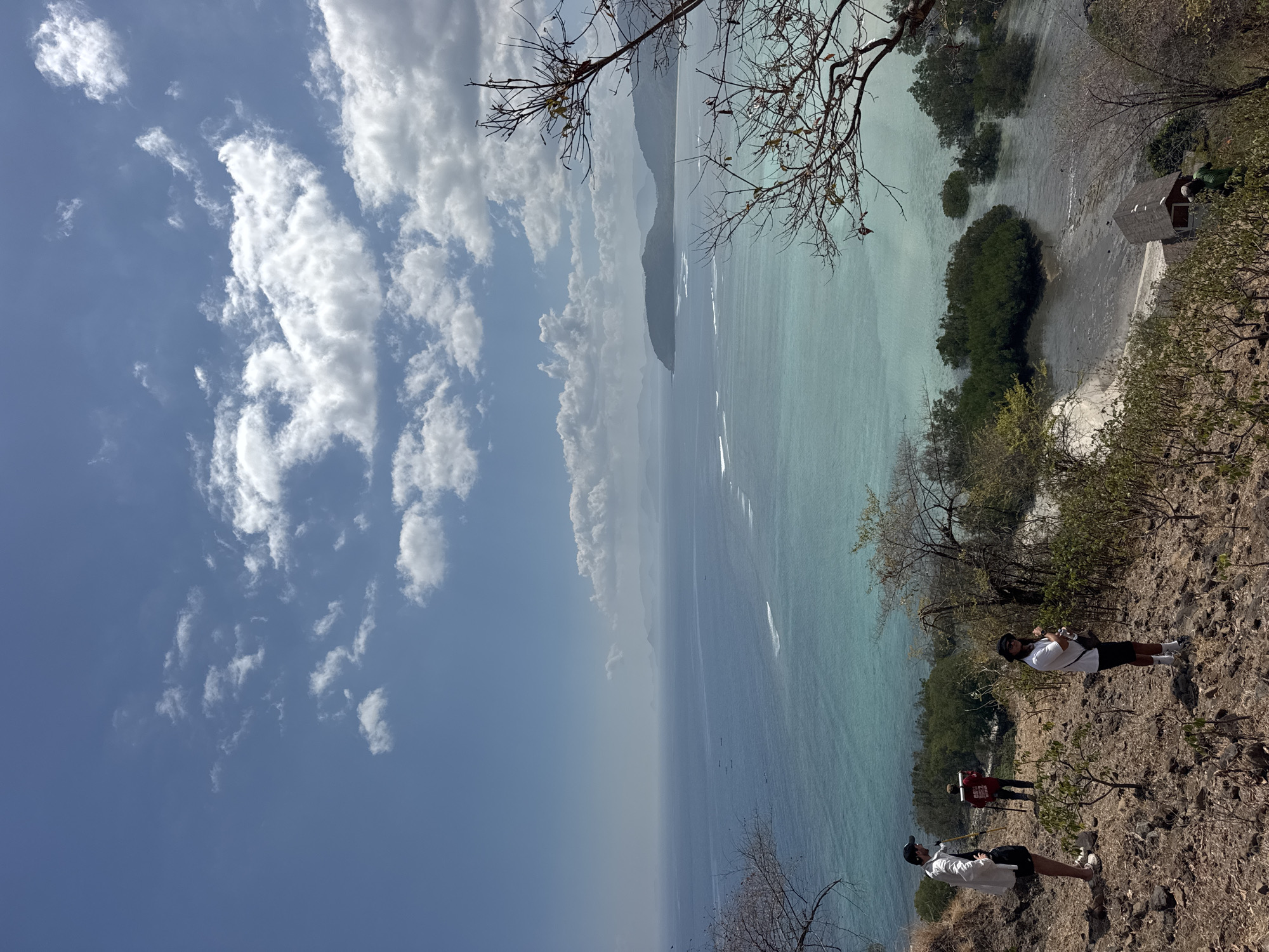 An elevated view captures a coastal landscape in Benete, Indonesia, featuring a wide expanse of the ocean under a partly cloudy blue sky. The water transitions from a clear turquoise near the shore to a deeper blue further out. Small boats are visible on the water.
In the foreground, a group of three individuals is present on a dry, rocky, and sparsely vegetated hillside. One person, wearing a white shirt and dark shorts, stands facing left with a dark hat and a stick. Another person, in a red shirt and dark pants, holds a stick and appears to be walking away from the viewer. The third individual, in a white shirt, dark shorts, and a hat, stands facing the viewer.
Further down the hillside, a small, dark-roofed building with red walls is visible near the water's edge. Mangrove trees grow in clusters in the shallow water surrounding the beach area. The overall environment suggests a natural, undeveloped coastal setting.