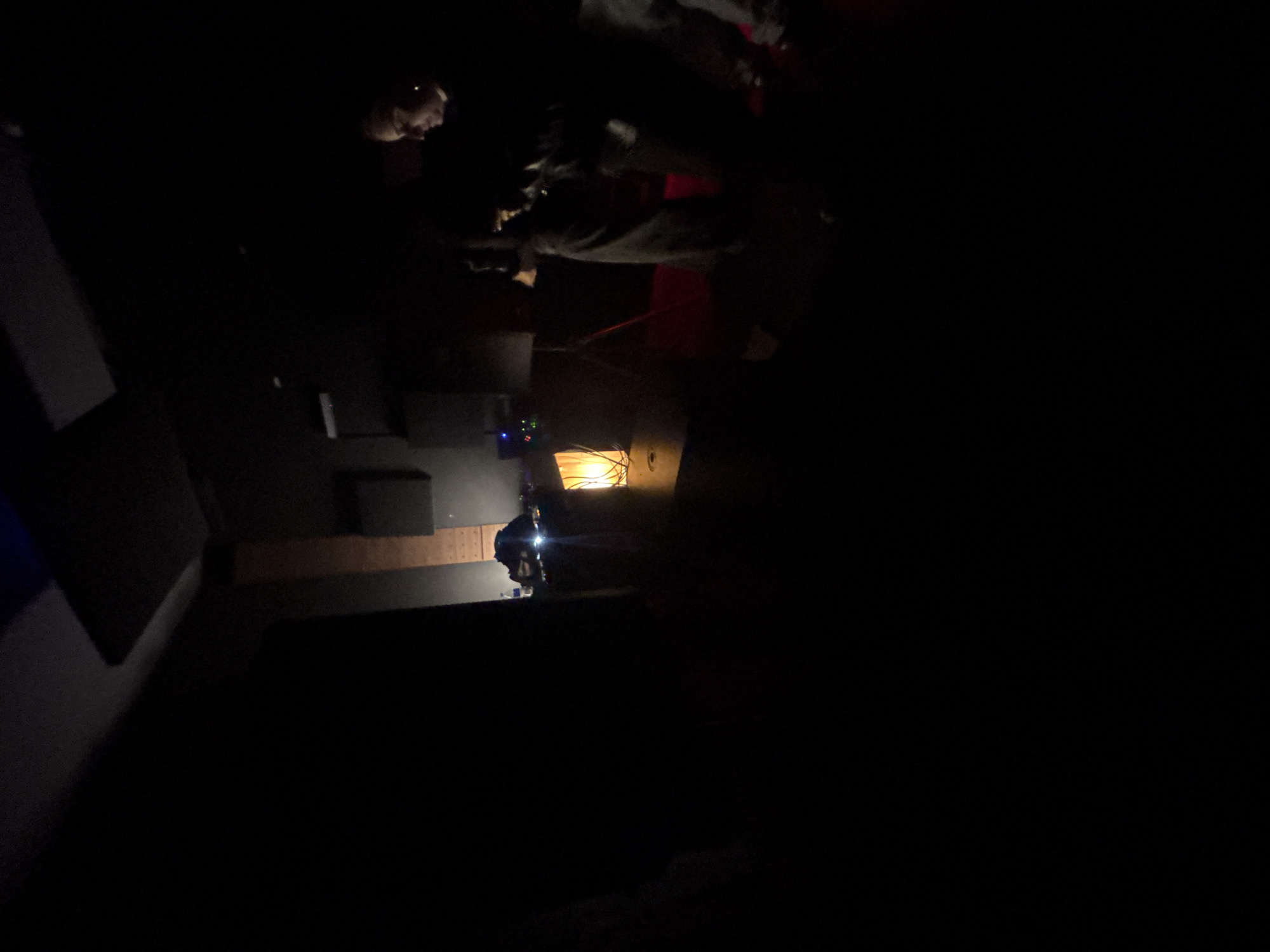 The image shows a dark indoor space, likely a recording studio. At the back of the studio, a person is seated behind a mixing console illuminated by a lamp. Another person is lying on the floor facing upward. To the side, another person is looking down. Equipment and cables are strewn about. The location and city of Buenos Aires, Argentina, cannot be verified in this image.