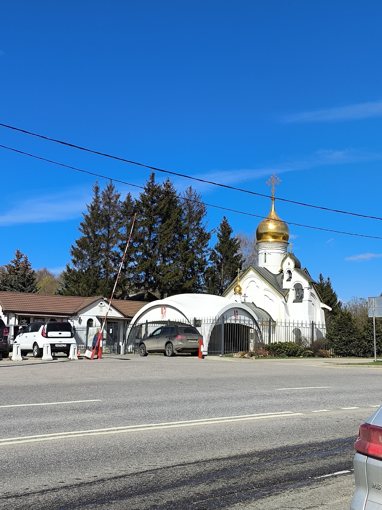 The image shows a Russian Orthodox church with a golden dome and a white exterior. The church is located behind a black metal fence and is surrounded by trees. In front of the church is a road with cars parked on the side. A red and white striped barrier arm is raised, likely controlling access to the property. The sky is blue with some clouds. The bottom of the image has the rear of a silver car. Without additional information, it is not possible to confirm the specific location in Moscow.