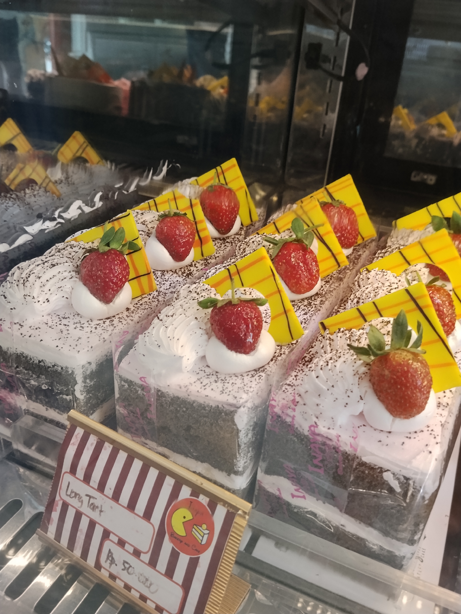 This image shows several slices of chocolate cake decorated with white frosting, black sprinkles, and fresh strawberries. Each slice also features a decorative yellow and orange triangular pastry garnish. The cakes are displayed in a refrigerated case, likely in a bakery or dessert shop. A price tag is visible in the foreground with the text "Long Tart" and "Rp. 50,000". The overall scene suggests a commercial food establishment. This bakery is located in Ciruas, Indonesia.