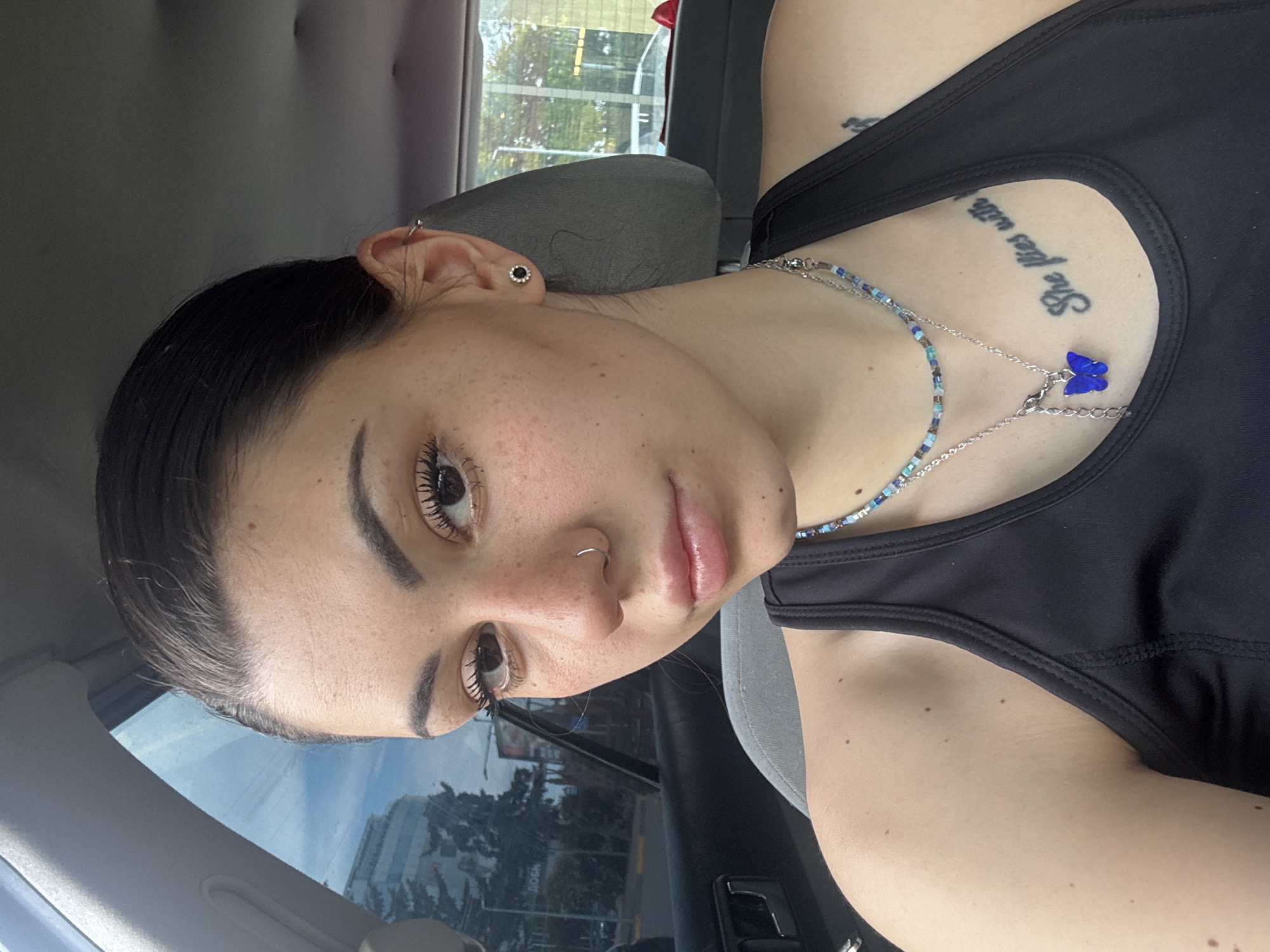 A close-up, portrait-oriented shot features a young woman inside a car, likely taking a selfie. She has dark hair pulled back, light skin, and visible freckles across her face, looking directly at the camera with a neutral, composed expression. She wears a black tank top or sports bra.
Her accessories include a silver hoop piercing in her nose, a small black stud earring in her earlobe, and another silver hoop in the upper cartilage of her ear. Two necklaces are visible around her neck: one a string of small blue, white, and silver beads, and the other a silver chain with a prominent blue butterfly charm. On her upper chest, a tattoo in a cursive script reads "She lives with."
The setting is the interior of a vehicle, with a grey headrest and parts of a car door visible. Through the car window, an outdoor scene is suggested, showing blurred trees and what appear to be buildings under bright daylight, indicative of daytime. The location context for this image is Sofia, Bulgaria.