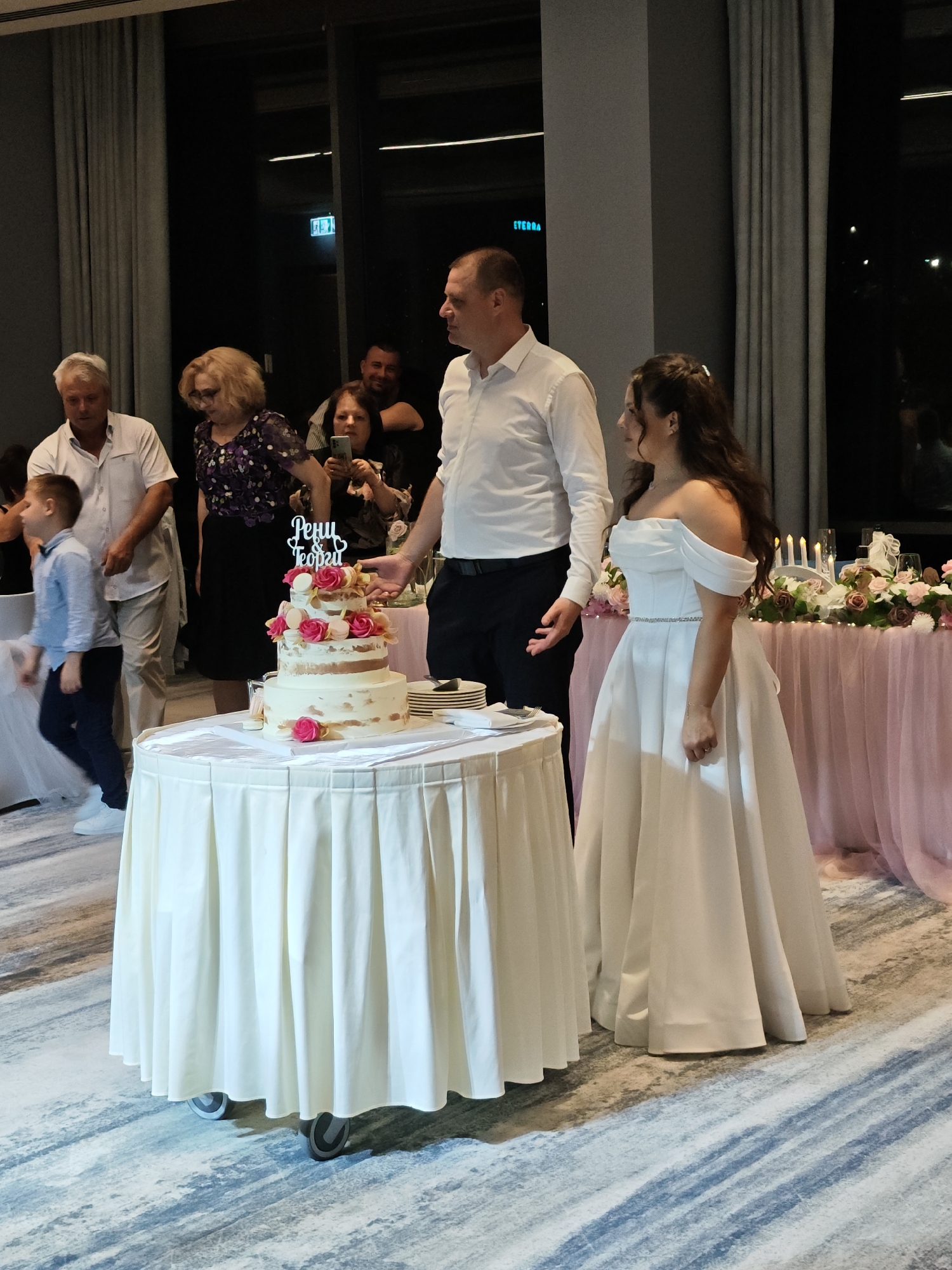 The image shows a wedding reception, likely taking place indoors. The couple, a bride in a white off-shoulder gown and a groom in a white shirt and black pants, stand next to a round table covered with a white cloth. On the table is a three-tiered cake decorated with roses and macarons, topped with a decorative topper, written in Bulgarian "Рени Георги", meaning "Reni Georgi". Plates are stacked nearby.
Several guests are visible in the background. To the left, an older man in a white shirt, a woman in a dark floral dress, and a young boy in a blue shirt are observing the couple. Another individual is partially visible, holding a smartphone, possibly taking pictures.
Behind the couple is a table draped with a pink cloth, adorned with candles, flowers, and other decorative items. Curtains are visible on the left side, and large windows showing a night scene are behind the couple. The scene is lit with artificial lighting. The floor is covered with a carpet with a gray-blue pattern. It's likely the location is in the city of Sofia, Bulgaria.