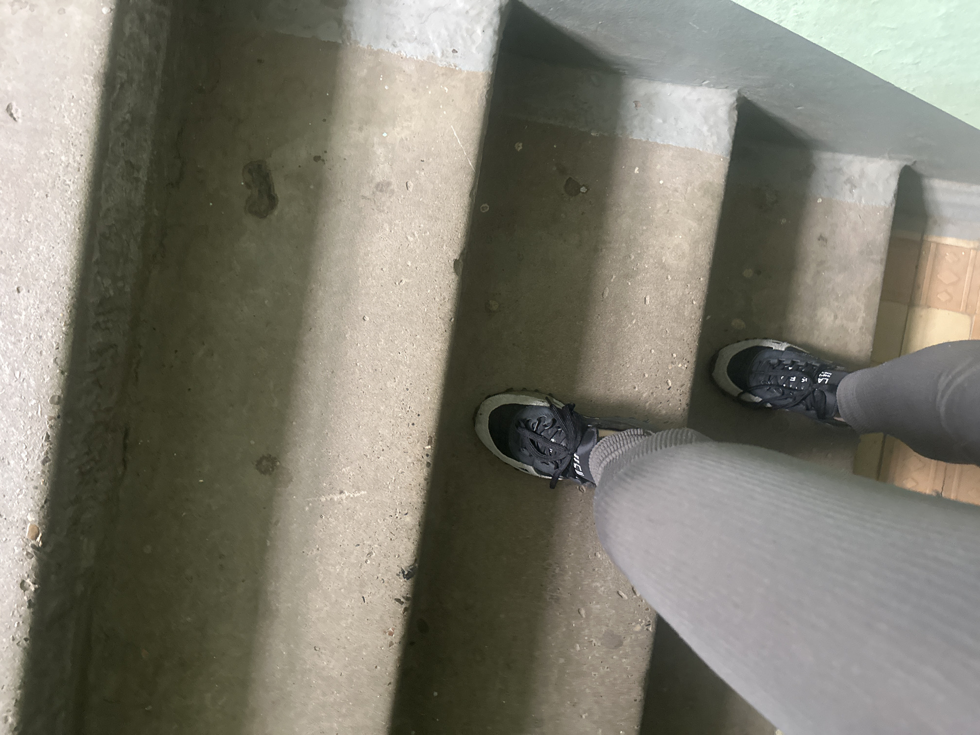 A downward-facing, high-angle shot shows a person standing on concrete stairs. The individual wears gray leggings and black running shoes with gray soles. The stairs are made of rough, textured concrete with gray paint on the top edges. The scene is indoors, possibly a stairwell. The location is unknown.