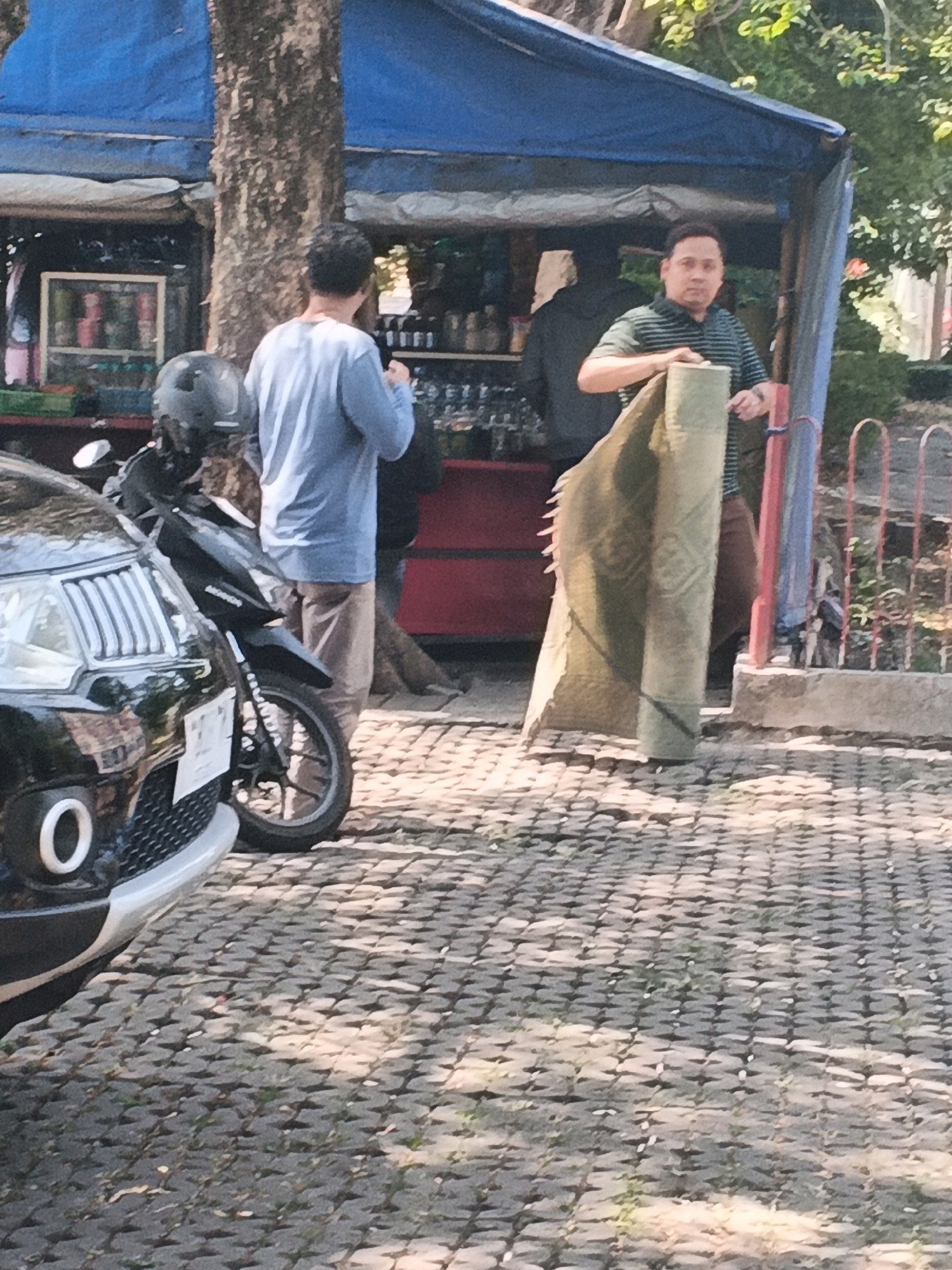 In Semarang, Indonesia, on a sunny day, a man is seen rolling up a patterned rug or carpet outside a stall. The stall has a blue tarp roof and displays various items behind a glass or plastic front. To the left, a black car and a black motorcycle are parked on a cobblestone-like paved area. Another person in a blue shirt and light-colored pants stands near the stall, facing away from the camera. The background shows a tree trunk and some foliage.