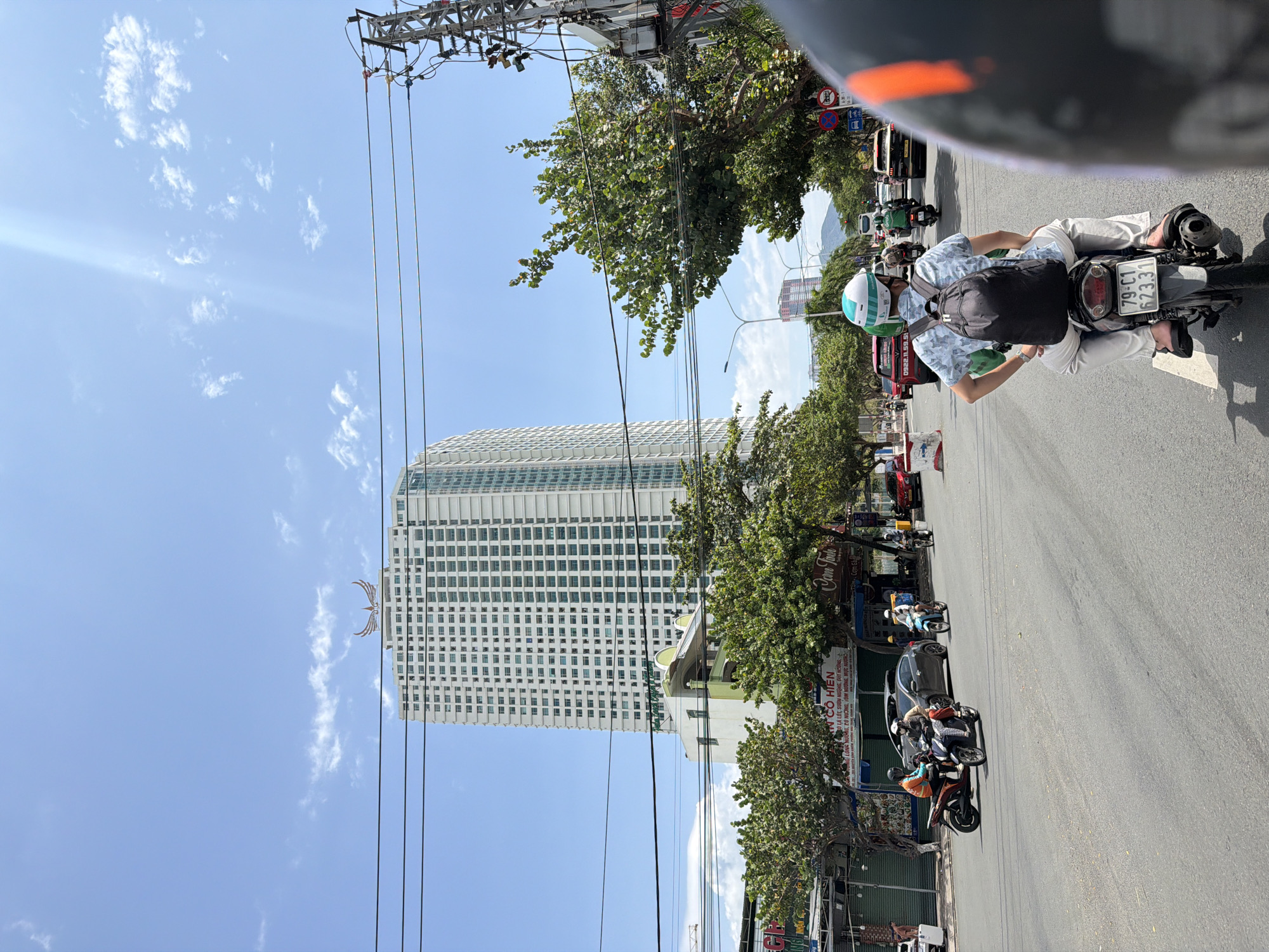 This image captures a vibrant street scene in Nha Trang Ward, Vietnam, on a bright, sunny day with a clear blue sky and scattered white clouds. The perspective is from a moving vehicle, likely a scooter, as indicated by the partial view of a helmet in the upper right.
In the immediate foreground, the back of a person riding a scooter is visible, wearing a light-colored, patterned short-sleeve shirt, a black backpack, and white pants, with a light green and white helmet. The scooter's license plate, "79-C1 623.31," is clearly visible.
The midground features a busy street with numerous scooters, a car, and a red bus, suggesting active traffic. Lush green trees line the street, partially obscuring storefronts and power lines. A dense network of overhead power lines and cables is prominent against the sky and above the trees.
In the background, a tall, modern high-rise building with a grid pattern of windows dominates the skyline. A decorative element, resembling wings or a crescent moon, is visible near the top of the building. Smaller buildings and distant mountains are also visible.
Visible text on a street-side sign reads "ẤN CÔ HIỆN" and appears to list menu items such as "Phở LÀ LỐT, SƯỜN NƯỚNG, CÚT NƯỚNG," and "TÔM NƯỚNG, MỰC NƯỚNG" (grilled shrimp, grilled squid), suggesting a restaurant or food stall. A phone number, "0922.11.59.51," is also displayed. The overall scene depicts a typical bustling urban environment in Vietnam.