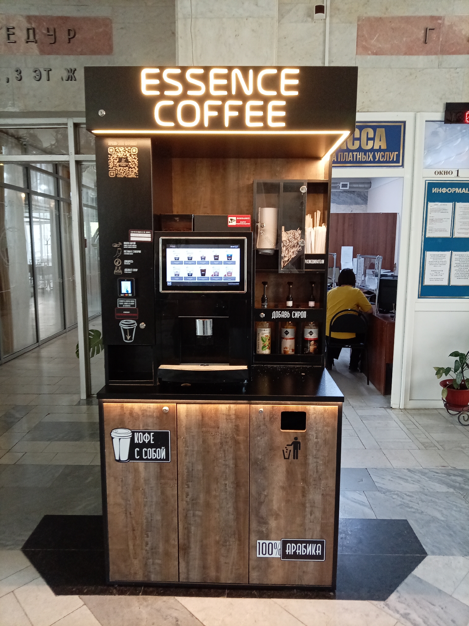 This is a full-shot, eye-level, indoor photograph of an "ESSENCE COFFEE" vending machine located in Zheleznovodsk, Russia. The machine is positioned in a hallway with a marbled floor and large glass-paneled doors to the left, suggesting it's in a public building such as an office or administrative center. Above and to the right of the coffee machine, signage indicates a "CASH DESK" and "WINDOW 1" for information, implying it's in a service area.
The coffee machine itself is predominantly black with illuminated white lettering reading "ESSENCE COFFEE" on its top facade. It features a touchscreen interface displaying various coffee options, a payment slot, and a dispenser. Below the machine, a wooden cabinet houses a trash receptacle on the right, and on the left, a sign with a coffee cup icon and Russian text "КОФЕ С СОБОЙ" (Coffee to go) is affixed. Another sign on the lower right of the cabinet reads "100% АРАБИКА" (100% Arabica).
On the right side of the image, a person wearing a yellow t-shirt and dark pants is seated at a desk, facing away from the camera, likely operating a computer. A potted plant is visible to the right of this individual. The background also shows architectural elements like columns and textured walls. The lighting appears to be a mix of ambient overhead light and the illuminated signage of the coffee machine.