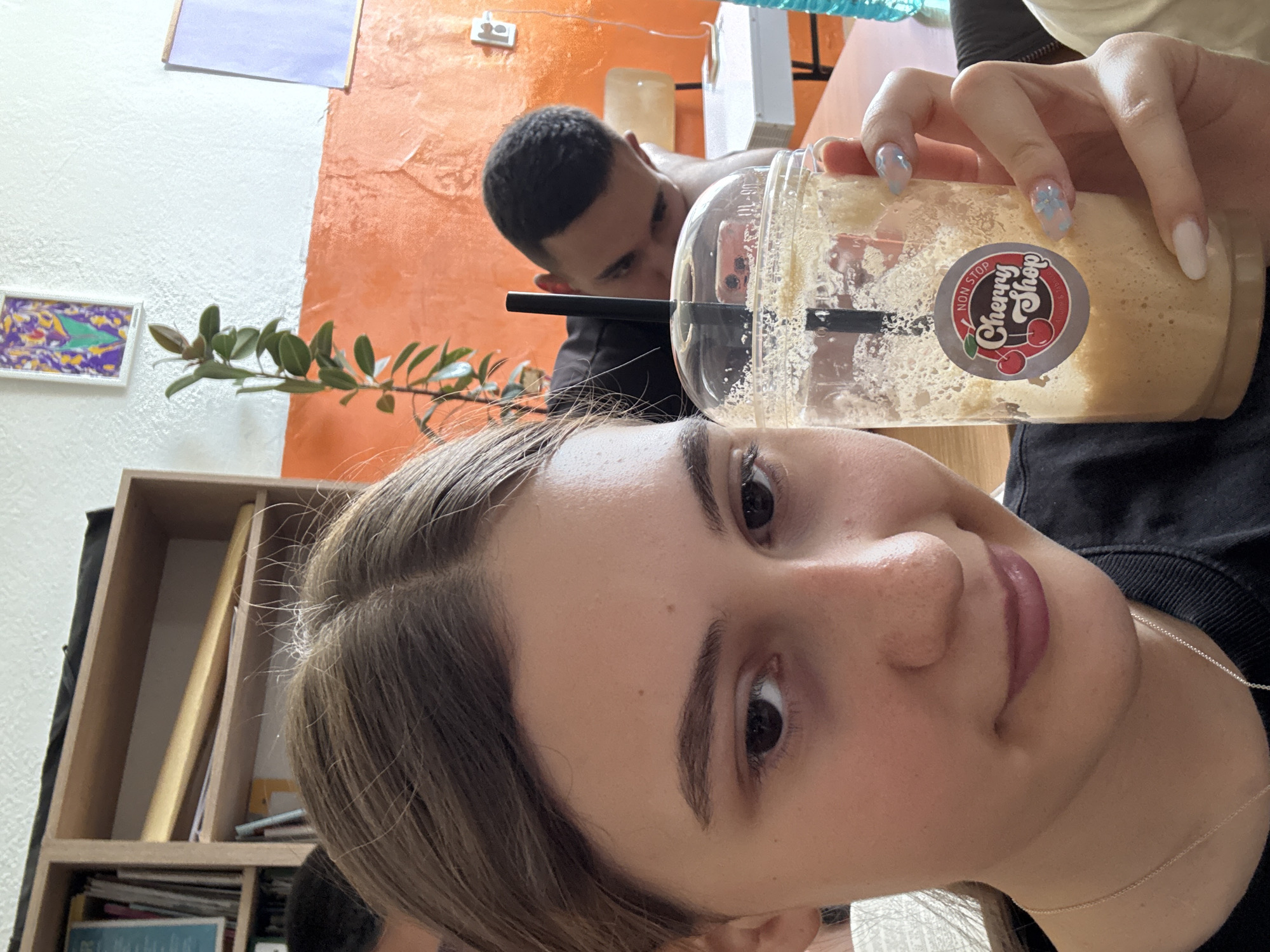 A selfie of a young woman inside an establishment. She is holding a partially consumed smoothie/drink from "Cherry Shop" in a clear plastic container with a black straw. In the background, a man peers curiously behind the drink. The location appears to be an interior setting with shelves, artwork, and a two-toned wall. No specific details link the location definitively to Panagyurishte, Bulgaria.