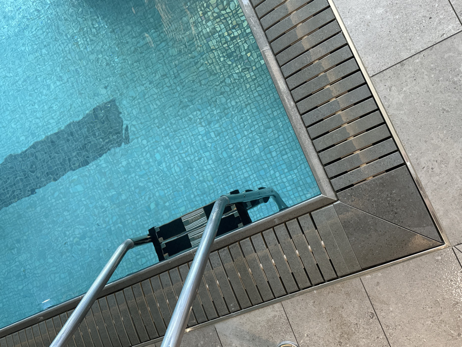 The image displays an overhead view of a section of an indoor swimming pool. The pool contains clear, turquoise water with visible surface ripples, revealing a mosaic pattern of small blue tiles on the pool floor. A stainless steel ladder with three black, textured anti-slip steps is partially submerged into the water, positioned at the pool's edge on the left. Surrounding the pool is a dark grey slatted drainage grate system, framed by a thin metallic strip. Adjacent to the grate, the pool deck is surfaced with light grey, large-format tiles. A triangular dark grey tile segment is visible at a corner where the slatted grate transitions to the main floor tiles. A long, dark, somewhat indistinct rectangular shadow is cast upon the pool bottom. No individuals or specific activities are depicted. The exact location within London, United Kingdom, cannot be determined from the provided visual information.