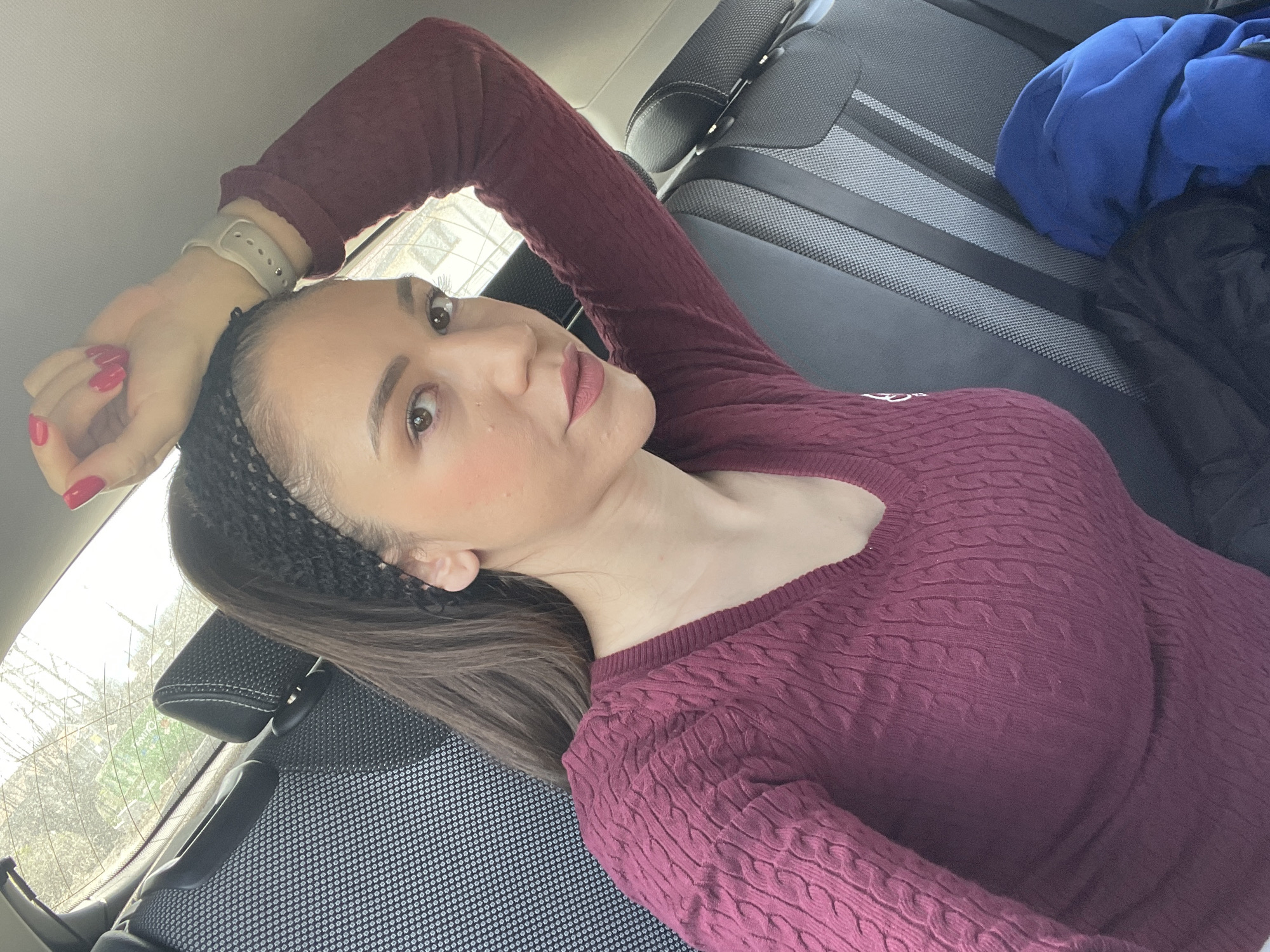 The image shows a woman lying down in the back seat of a car. She is wearing a maroon-colored sweater and a black headband. Her hair is brown and pulled back. She is looking directly at the camera with a slight smile. Her right arm is raised above her head, and she is wearing a watch on her wrist. Her nails are painted red.
In the background, the car seat is visible, upholstered in gray and black fabric. There is a blue garment and a dark garment partially visible on the seat next to her. Outside the car window, there are trees with bare branches.
There is insufficient information to determine if the location is Dobrogled, Bulgaria.