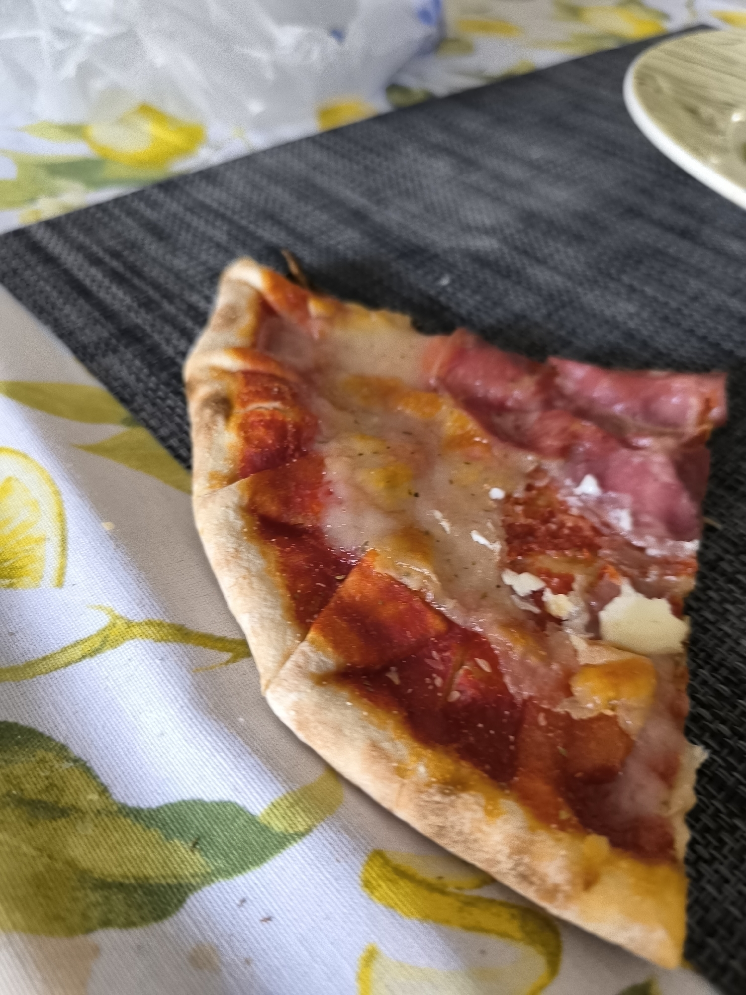 The image shows a partially eaten slice of pizza resting on a white tablecloth patterned with lemons. The pizza has a golden-brown crust, a red tomato sauce base, and toppings including what appears to be salami and mozzarella cheese. The slice sits on a black woven placemat, near a white plate. The location is unknown and cannot be determined from the image. There are no people visible. The setting appears to be an indoor dining environment.