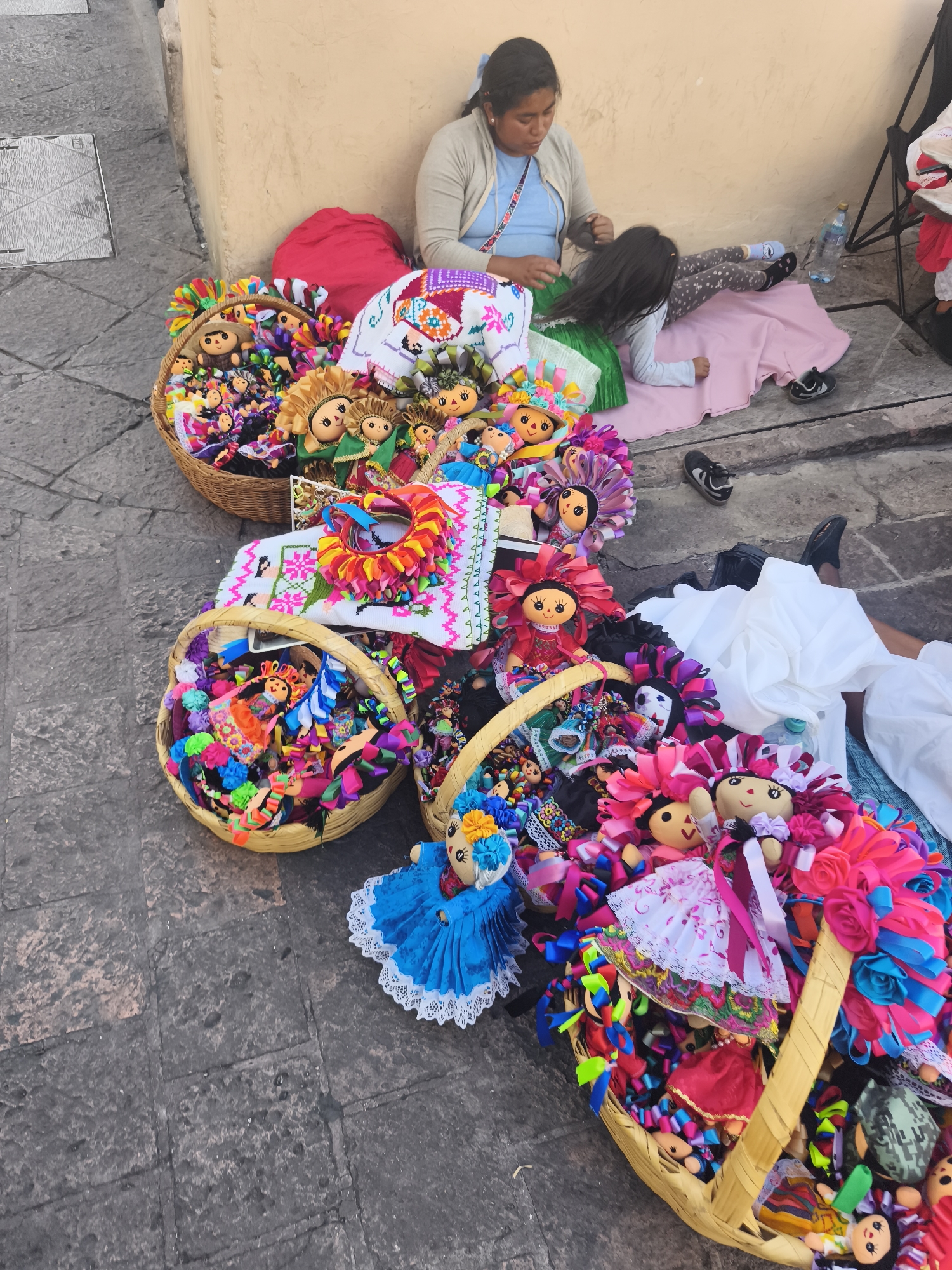 The image depicts a street scene in Querétaro, Mexico. A woman with dark hair is sitting against a yellow wall, braiding the hair of a young girl lying on a pink blanket. Several woven baskets filled with colorful, handmade "María" dolls are scattered around them on the cobblestone pavement. A red pillow and embroidered textile are also visible among the dolls. The scene appears to be a marketplace or street vending setup, with the woman likely selling the dolls. A few other people are partially visible in the background. A black and silver utility hatch is visible on the ground to the left of the image.