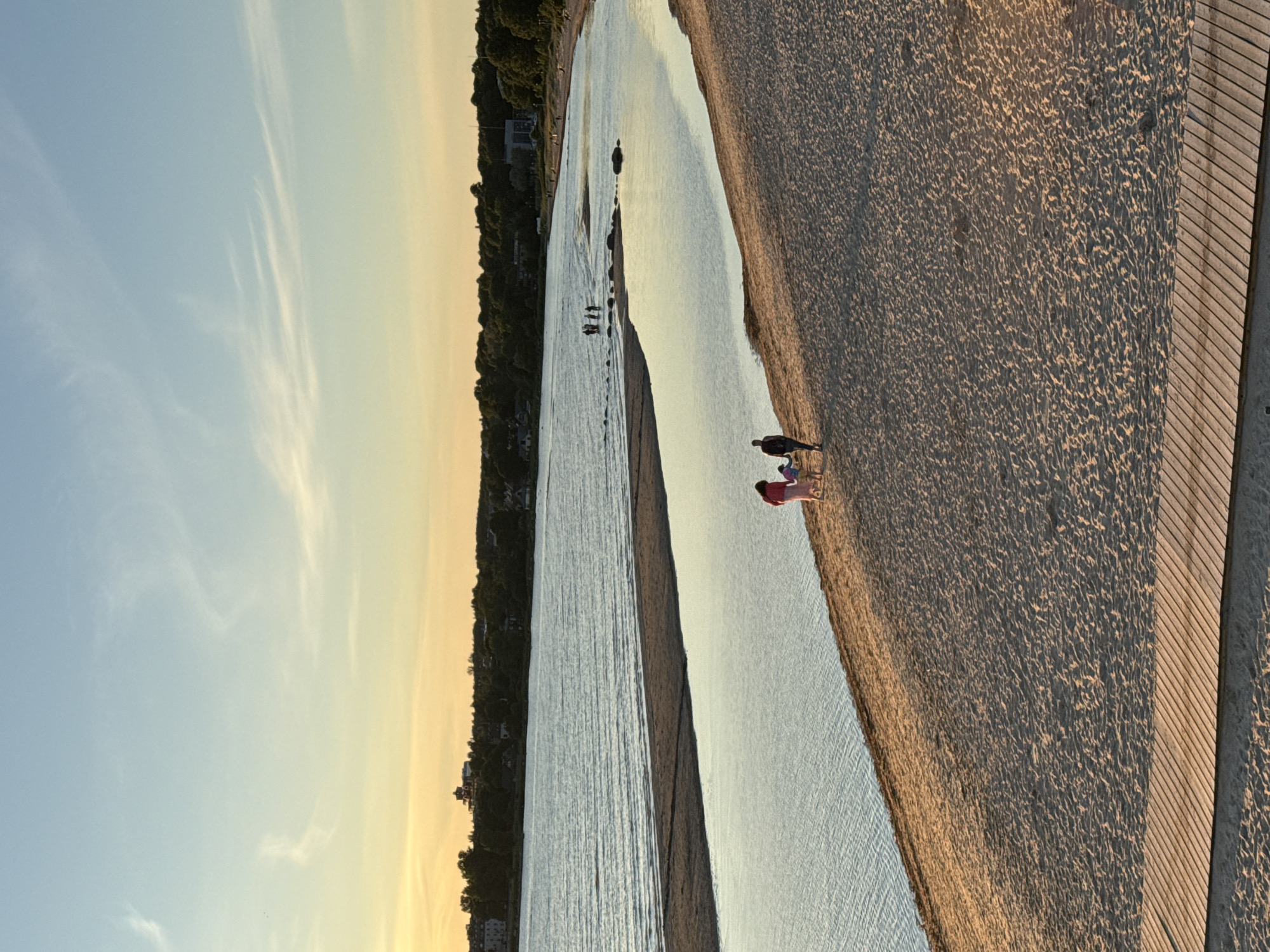 In the city of Tallinn, Estonia, a beach scene unfolds at sunset. The sky transitions from a pale yellow near the horizon to a soft blue overhead, with thin white clouds streaking across. The beach itself is composed of fine, light brown sand, marked with rippled textures. A wooden boardwalk runs along the bottom of the frame, adding a geometric element to the natural landscape.
The water of the sea is calm, reflecting the hues of the sky. A narrow sandbar separates a shallow, glassy inlet from the main body of water. Several people are visible in the distance, walking along the water's edge.
In the foreground, on the sandy beach, a small group of three people walks away from the camera. The person in the middle, a child, holds hands with the two adults on either side. The adult on the left wears a bright pink garment, while the adult on the right wears dark clothing. The lighting suggests it is late afternoon or early evening, casting long shadows. In the background, a dense line of green trees forms a backdrop to the shoreline, with buildings and structures visible amongst them, suggesting proximity to urban areas.