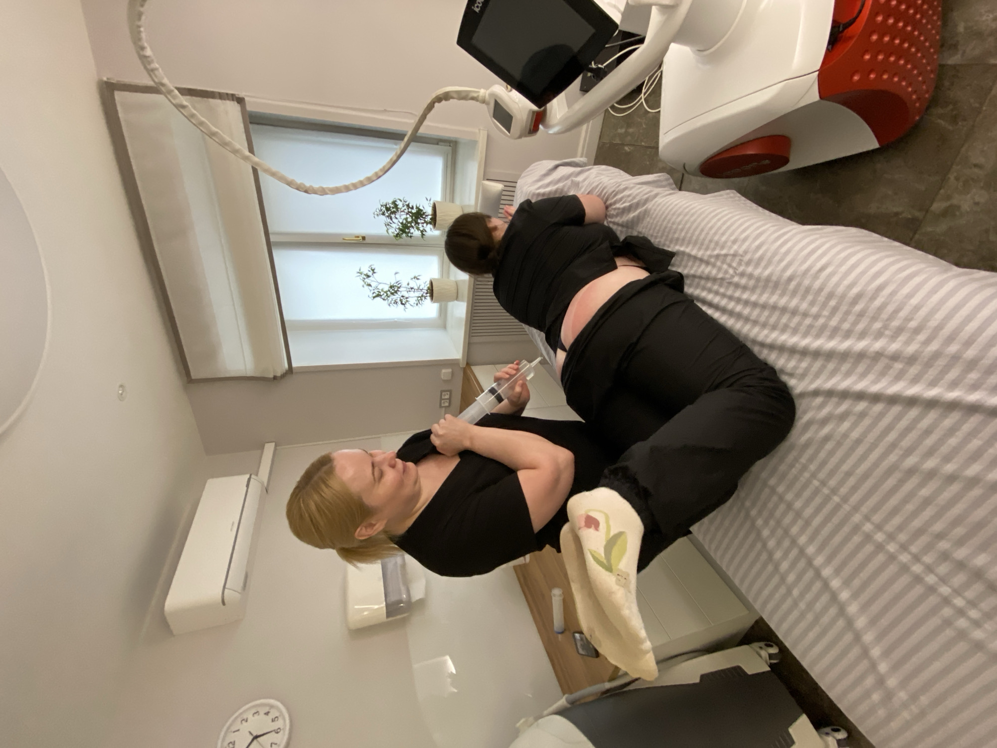 A healthcare professional in black attire is injecting a solution into a patient's lower back, while the patient lies face-down on a striped treatment table. A medical device with a display screen is positioned near the patient's head. The scene is indoors, with natural light entering through a window. Other medical equipment is visible in the background, including a small air conditioner or heater on the wall and a medical cart next to the treatment table. A clock is partially visible on the wall, showing the time as past 10:30. The location is inferred to be a medical or cosmetic treatment room, with the city and country indicated as Votkinsk, Russia.