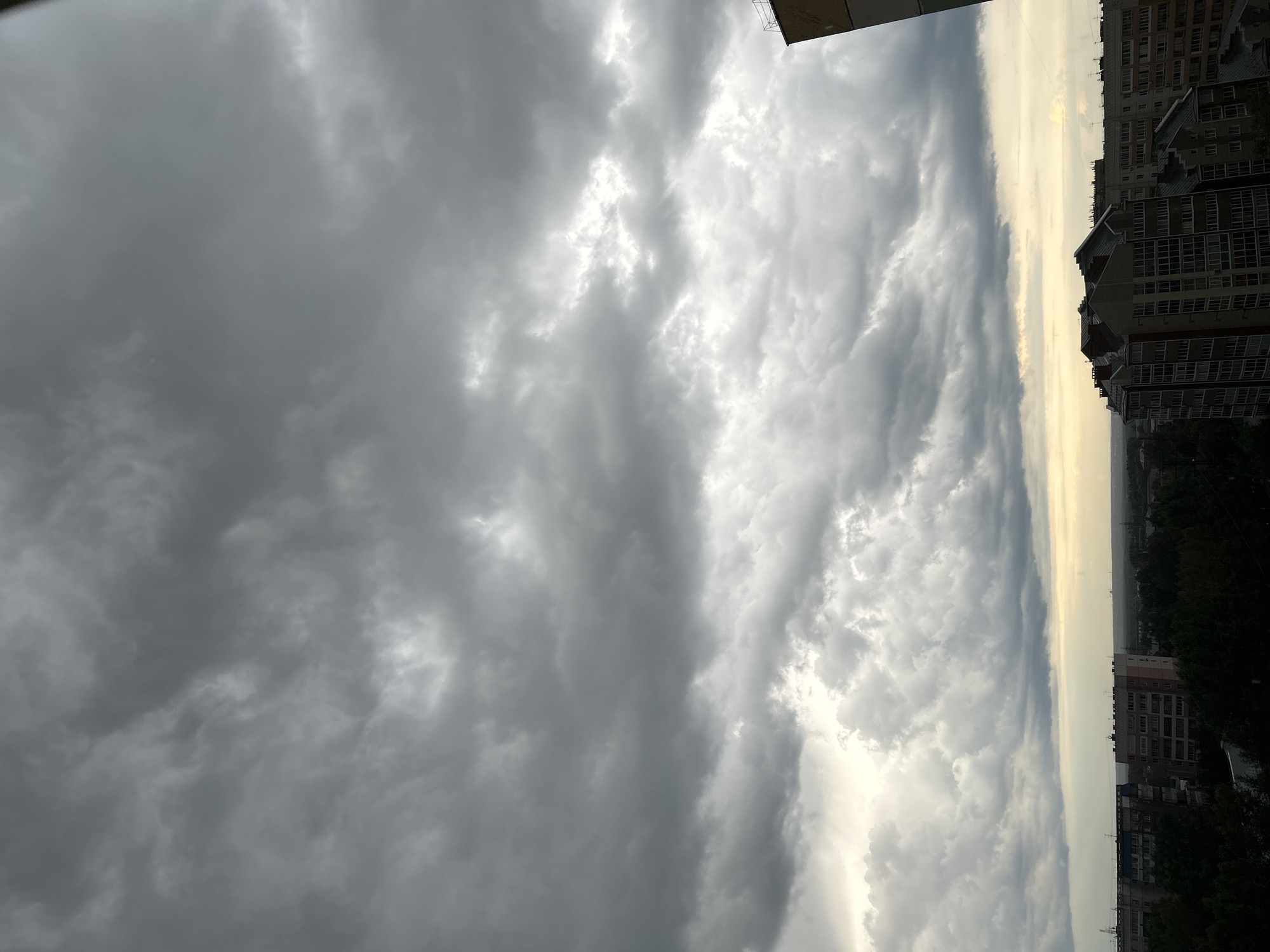 This is an outdoor, eye-level, horizontal shot of a cityscape under a heavy cloud cover. The sky is dominated by dark gray clouds with some lighter patches suggesting breaks in the cloud cover. The lower portion of the image shows a dense urban environment with several multi-story apartment buildings, some with visible windows and balconies. Green trees are interspersed between the buildings and in the foreground. A sliver of hazy yellow-orange light is visible on the horizon, indicating the direction of the sun, likely setting or rising. The scene depicts an overcast day in the city of Tomsk, Russia. There are no people visible in the image.