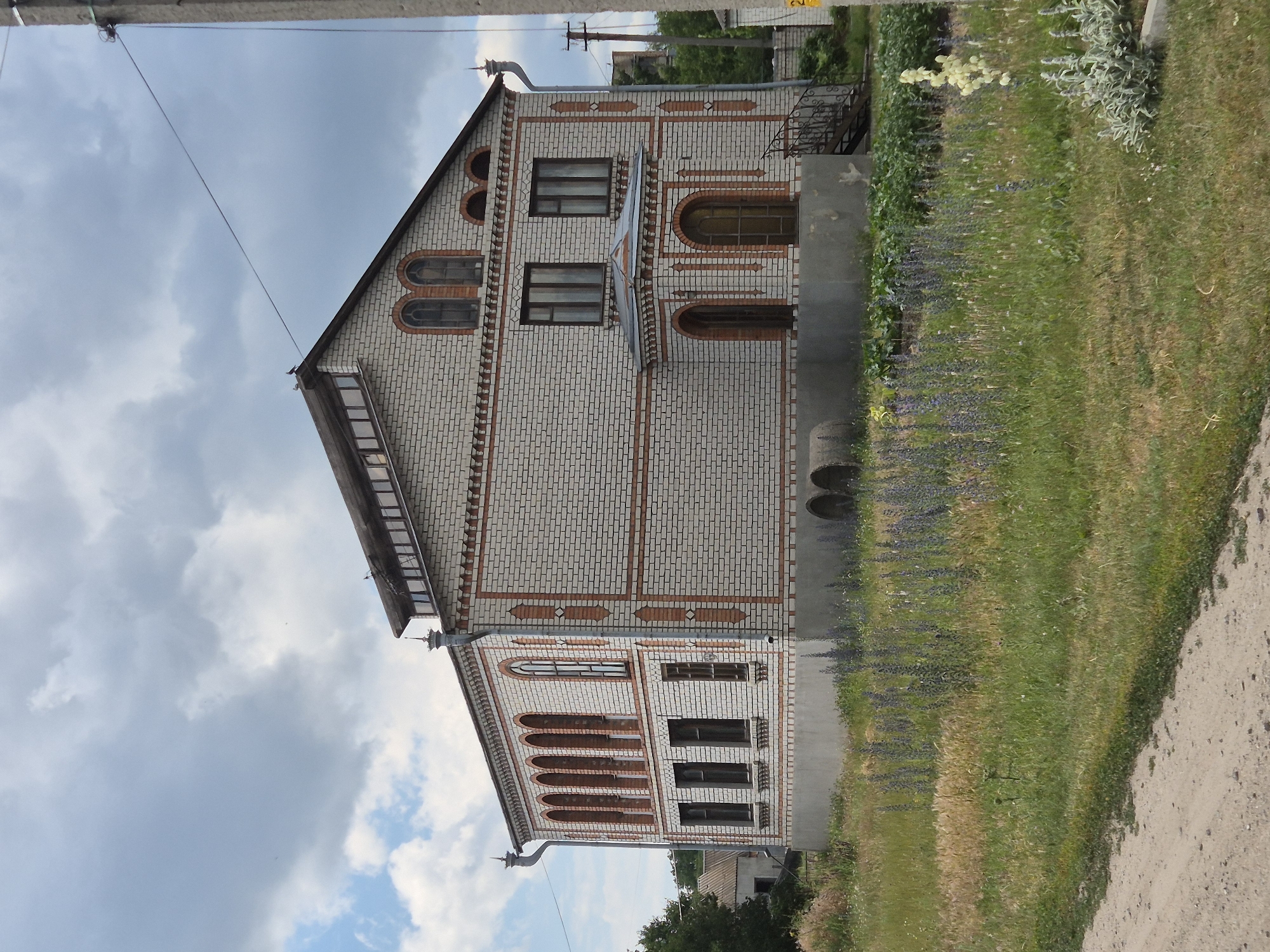 The image shows a two-story brick building with an ornate facade, located in the village of Sozonivka, Ukraine. The building is constructed of light-colored bricks with decorative red brick accents around the windows and corners. The roof is dark and sloped, with a row of windows along the top story under the eaves. The building has a foundation that appears to be made of concrete.
In front of the building, there is a grassy area with patches of wildflowers and some taller plants. A dirt path runs along the left side of the image. In the background, to the left of the building, another smaller building with a lighter roof is visible, and trees are present. To the right of the main building, a utility pole with wires extends upwards, and a section of a white fence is also visible. The sky is cloudy, with patches of blue sky peeking through.
There are no people visible in the image. The scene appears to be a quiet, rural setting. The building's architecture suggests it might have been a public building, such as a school or a cultural center, or possibly a large private residence.