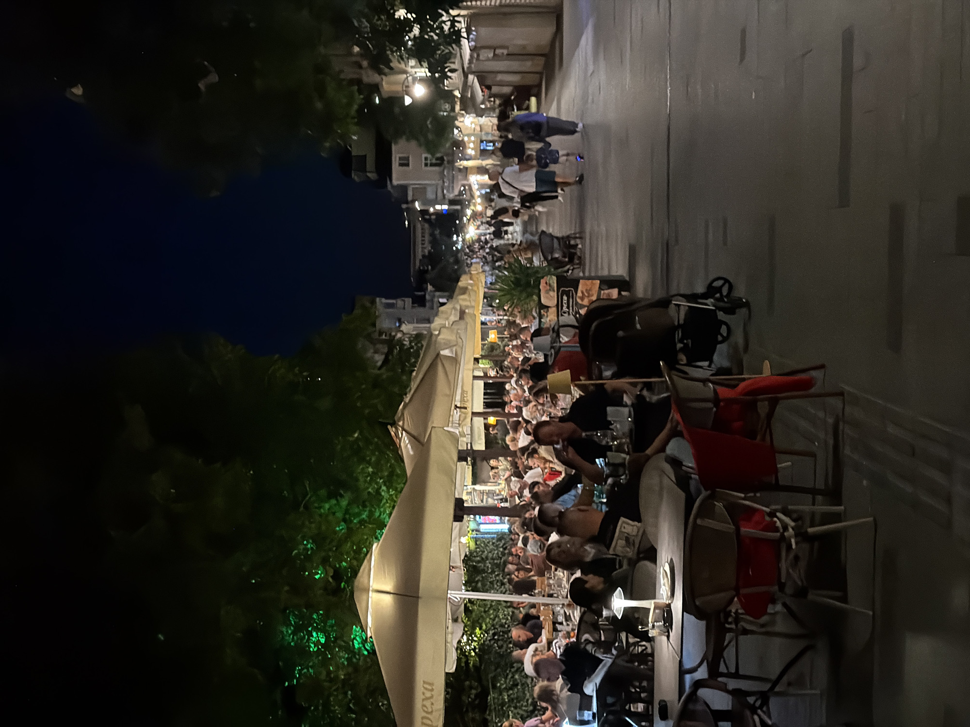 The image depicts a lively night scene on a pedestrian street in Burgas, Bulgaria, likely the pedestrian area along Bogoridi Boulevard. The scene is filled with people dining outdoors at restaurants and cafes. Umbrellas provide shade over tables where patrons are seated. In the foreground, several tables and chairs are visible, some with red cushions or jackets draped over them. A table lamp illuminates one table. Further down the street, a family walks, with a child holding a parent's hand. Other individuals are seen strolling along the street. Buildings line the sides of the street, and trees with green lighting contribute to the ambiance. The overall impression is one of a bustling urban environment at night, with a focus on outdoor dining and leisurely evening activity.