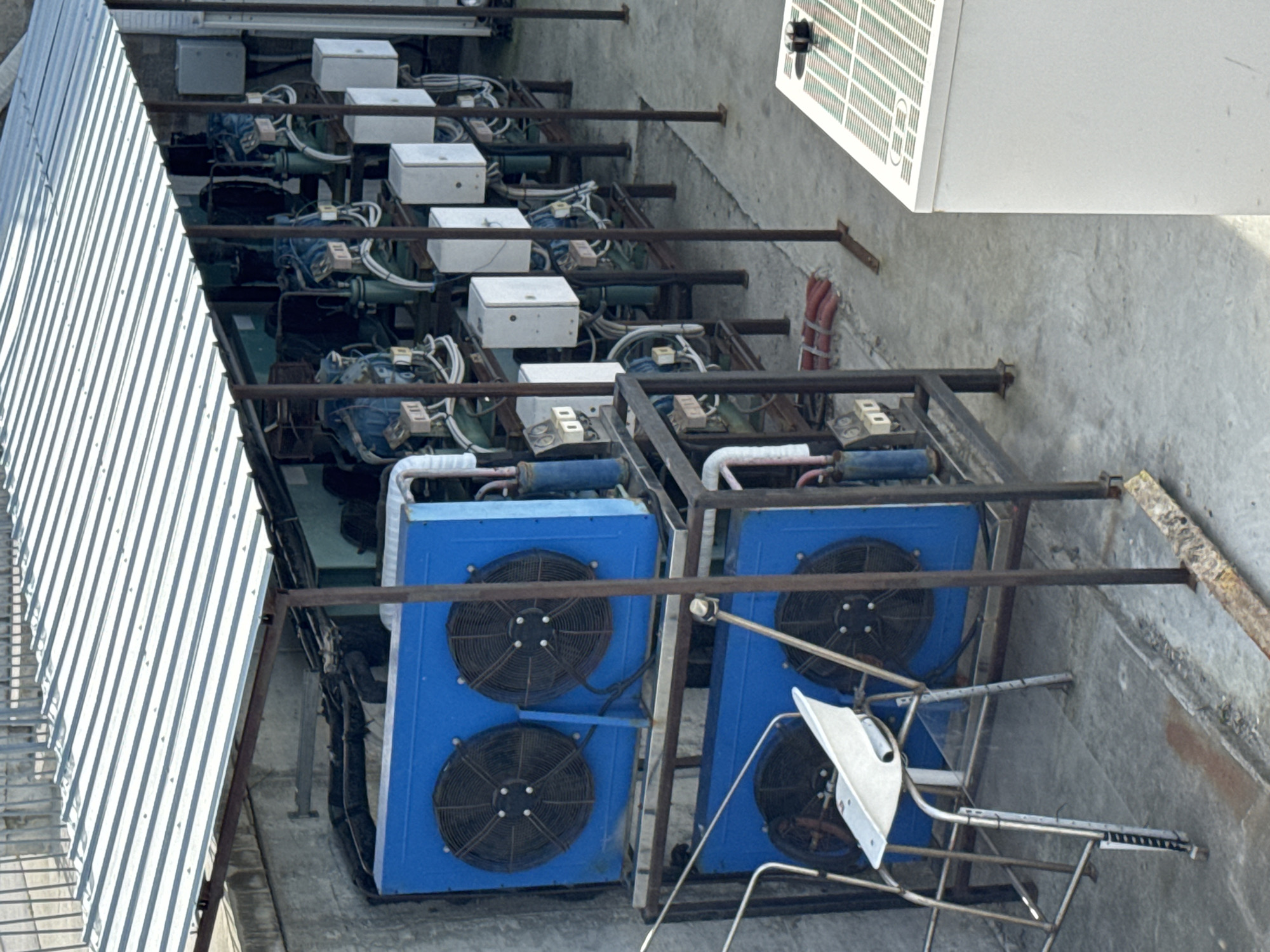 The image displays an extensive array of industrial machinery situated outdoors on a concrete surface, likely a rooftop or an elevated service platform, viewed from an overhead perspective. The original image, oriented vertically, shows the machinery stretching from the upper-left to the lower-right.
Along the lower-right side of the frame, two prominent, rectangular blue units are visible. Each unit features at least two circular black fan grilles, indicative of condenser units or chillers, possibly part of an HVAC or refrigeration system. These units are mounted on a robust, dark brown metal framework that supports the entire installation.
To the upper-left of these blue units, the machinery extends in a long row, comprising multiple sections. Each section includes large, dark green or blue components, likely compressors, along with several smaller white rectangular boxes that appear to be electrical control or junction boxes. A dense network of pipes, conduits, and cables intricately connects all these components, suggesting a complex operational system.
To the far upper-left of the image, a sloped, corrugated silver metal roof or wall of an adjacent building is prominently featured. The ground beneath the machinery is a uniform grey concrete slab. In the upper-right background, a partially obscured white rectangular panel with a grid pattern is visible. Bundles of red-wrapped cables are neatly arranged in the upper-middle section. In the lower-right foreground, a small, irregularly shaped, light-colored metallic object rests on a slender metal stand.
The scene is brightly lit by daylight, indicating clear weather conditions. There are no individuals present, and no specific activity is taking place; the machinery appears static and functional. No discernible text is visible on any of the equipment. This setting suggests a utility or service area of a commercial or industrial facility, located in Varna, Bulgaria.