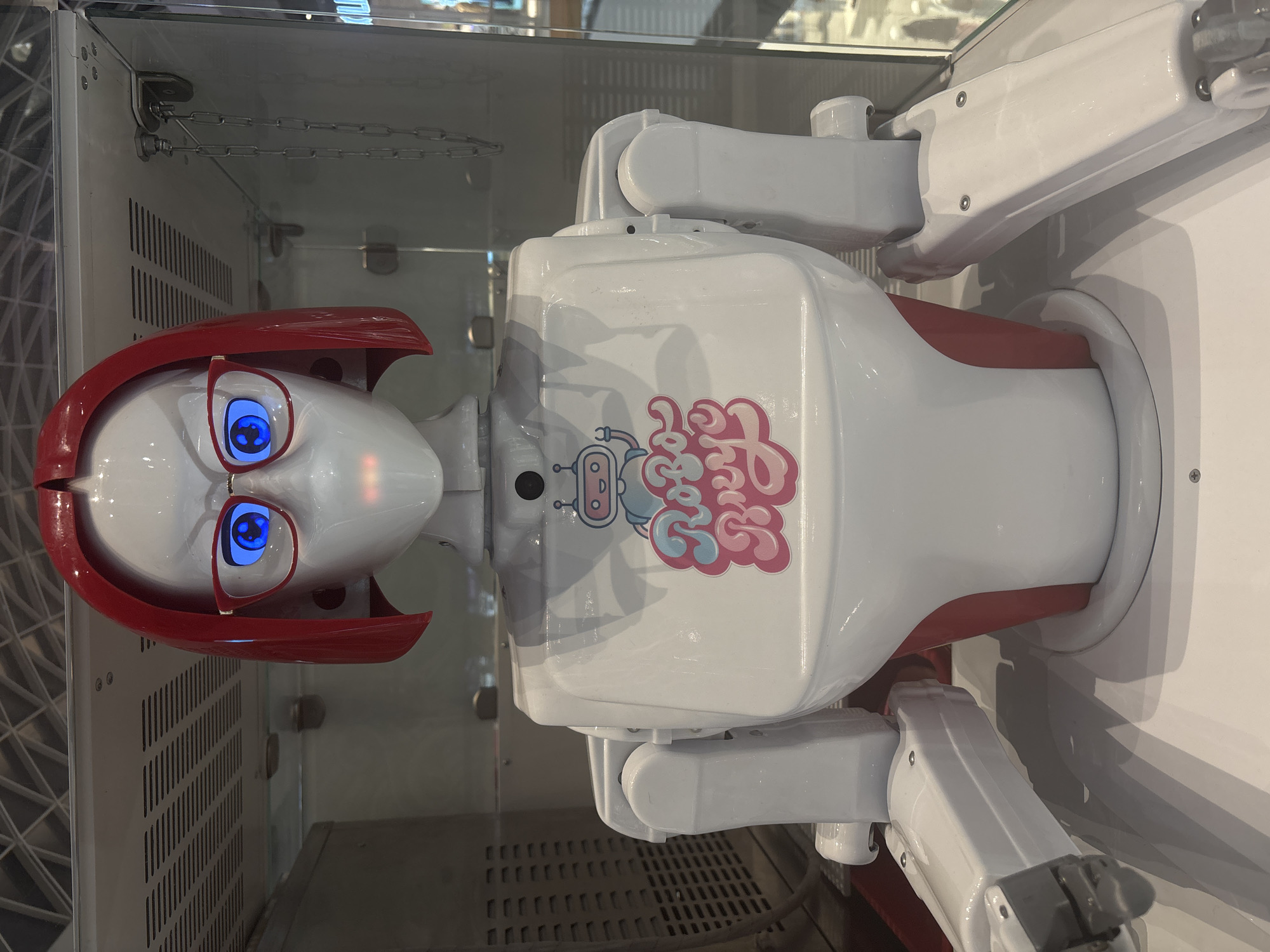 An upright, white humanoid robot with prominent red and pink details is displayed within a clear glass or acrylic enclosure. The robot's head features a smooth white face, vibrant glowing blue eyes with circular pupils, and red-framed glasses. A stylized red bob-cut headpiece or hair covers the top and sides of its head. Its torso is largely white, complemented by a reddish-pink panel underneath. On its chest, a distinctive logo is visible, composed of a small, light blue and pink cartoon robot next to the words "Funky Fresh" rendered in a playful, bubbly font, predominantly in pink hues.
The robot's white, jointed arms extend downwards from its shoulders. Inside the display case, background elements include metallic surfaces, some with ventilation-like grates, and visible chains secured to the upper structure. The illuminated blue eyes suggest the robot is powered on. The setting appears to be an indoor environment, such as a museum, science exhibit, or retail space, in Perm, Russia, showcasing the robot as an object of interest or a functional display.