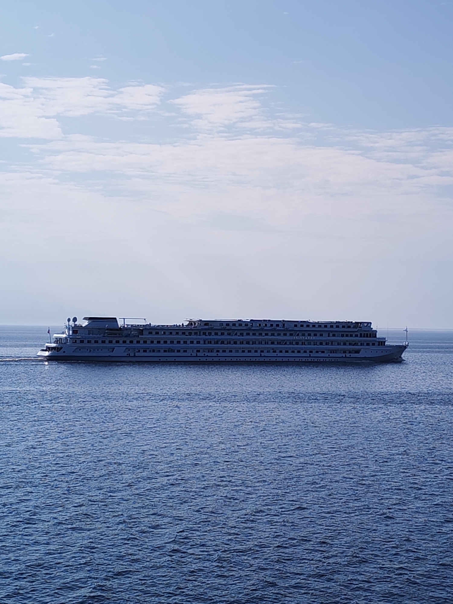 A large cruise ship navigates a body of water under a partly cloudy sky. The ship is white with multiple decks and numerous windows. The water is dark blue with small ripples. The environment appears to be a calm, open space with the horizon visible in the background. The location is Bol'shie Mkhi, Russia.