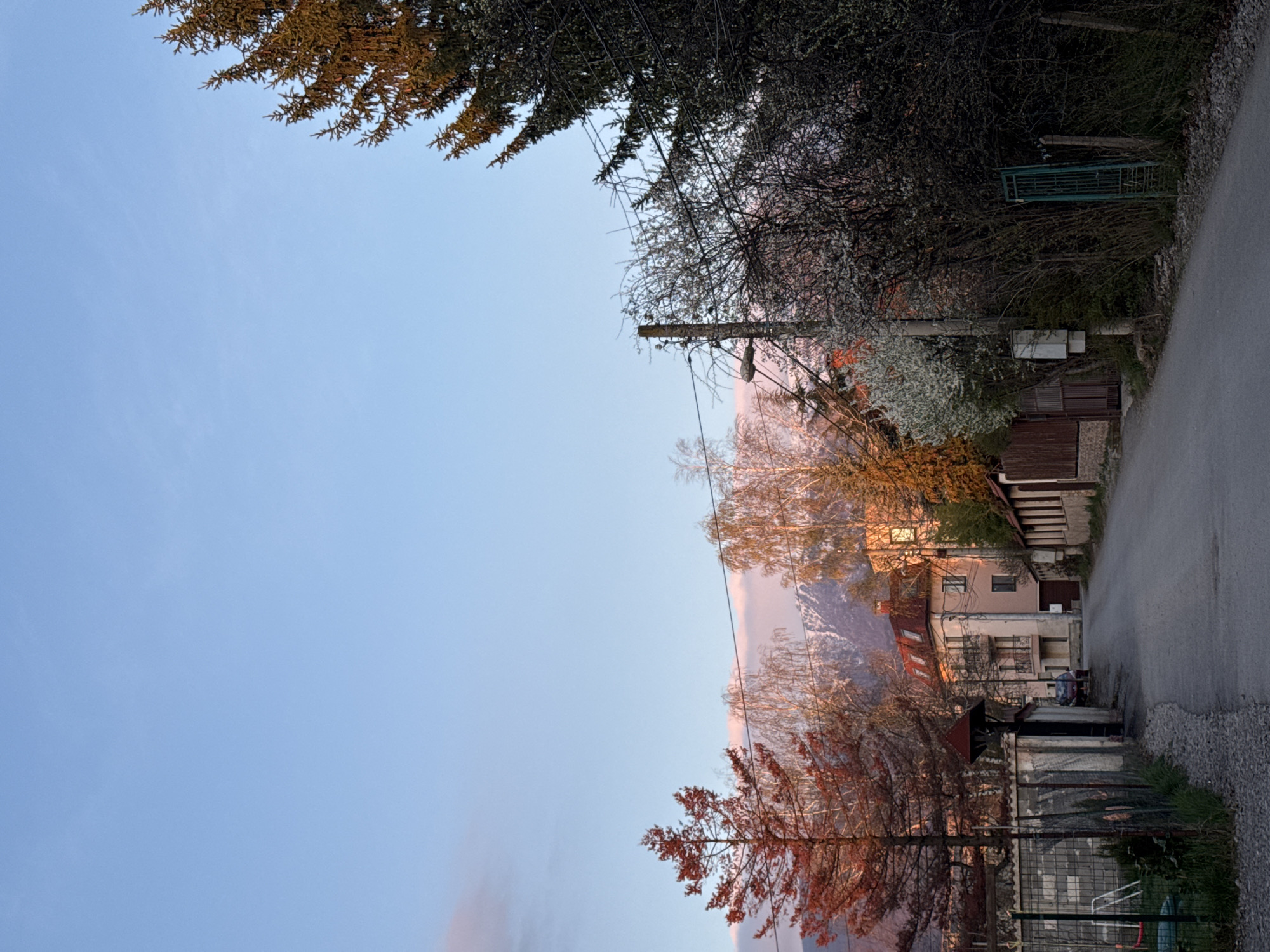 An outdoor scene shows an asphalt road on the right side, with a gravel shoulder. Vegetation lines the right edge, leading to a house with a fence and gate. Utility poles and wires are overhead. On the left, there is a clear blue sky with trees framing the top corners. In the center, there is a mountain visible in the background with light pink clouds on the horizon. The location may be in the city of Sofia, Bulgaria.