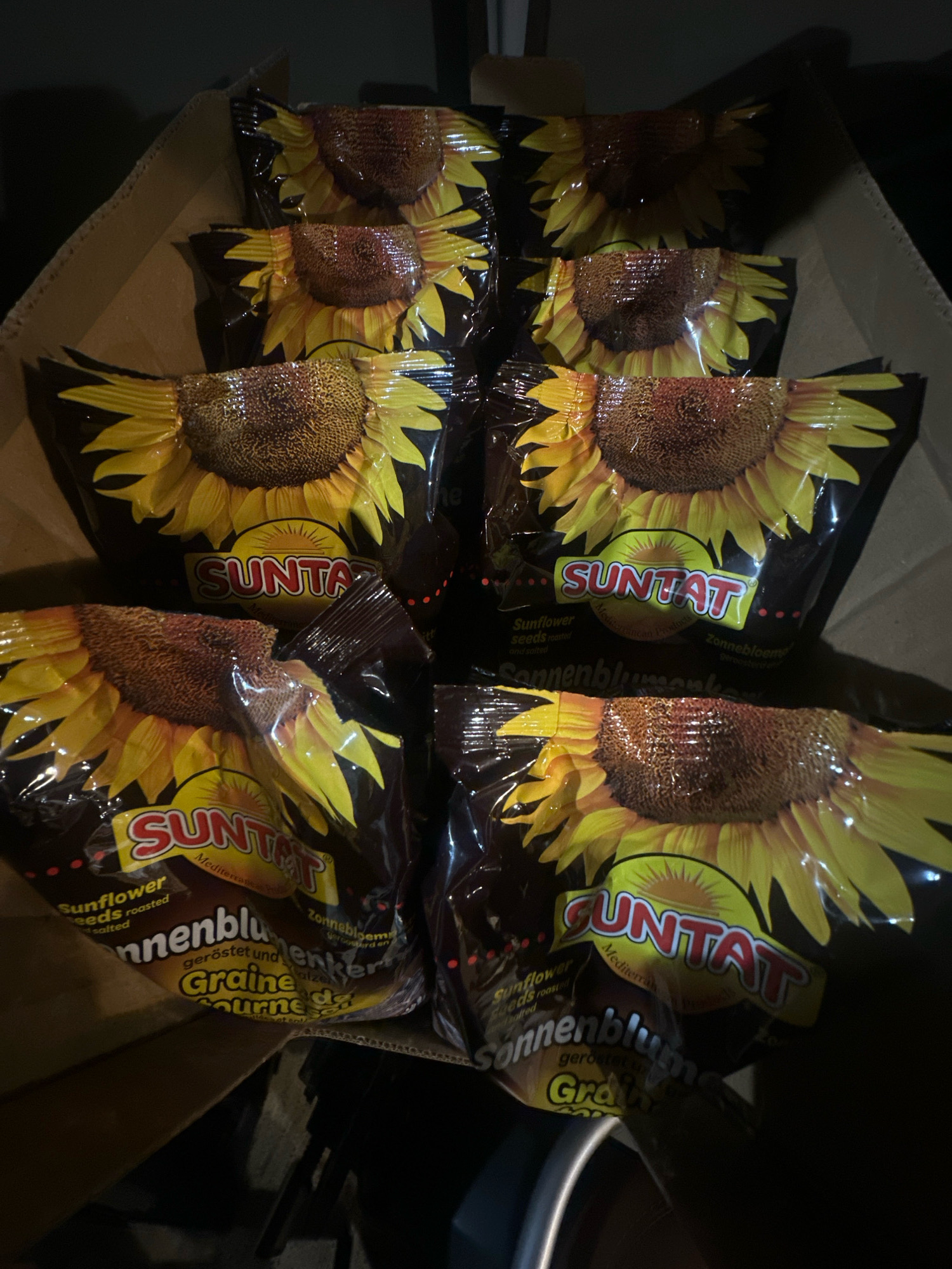 This image, taken in the city of Lapės, Lithuania, depicts a cardboard box filled with multiple packets of "SUNTAT" brand sunflower seeds. The packets feature a large sunflower image and branding in yellow and red. The box is partially open, revealing stacks of these seed packets. The lighting is dim, suggesting the photo was taken indoors in a low-light environment. No people are visible in the image.
