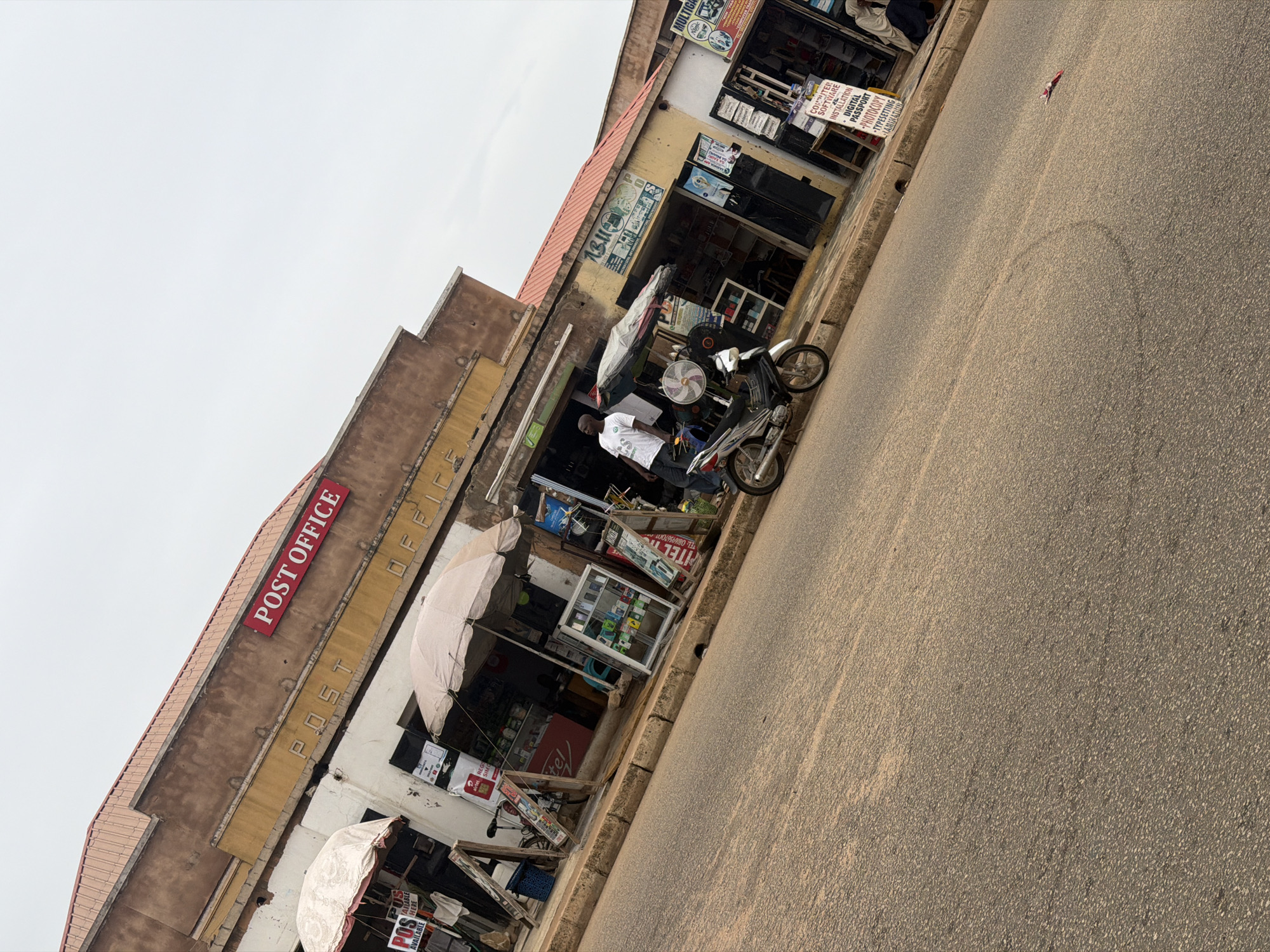 The image shows a street scene in Bukuru, Nigeria, with a Post Office building prominently featured. The Post Office sign is red and white, situated above the entrance. In front of the Post Office, several small businesses operate under makeshift awnings. To the left, a stall displays "POS AVAILABLE HERE" signage, and a bicycle is parked nearby. Further down, a beige umbrella shades a stall filled with various goods, including a display case of what appear to be mobile phones or electronic accessories. A motorcycle is parked in front of another stall, which has advertisements for phone services and accessories. A man in a white t-shirt is standing near the motorcycle. To the right, another stall has a sign advertising "COMPUTER SOFTWARE INSTALLATION DIGITAL PASSPORT PHOTOCOPYY TYPESETTING LAMINATIN," and a person in traditional clothing is seated nearby. The street is paved with asphalt, and the weather appears overcast. The buildings have a weathered facade, typical of older structures in the region.