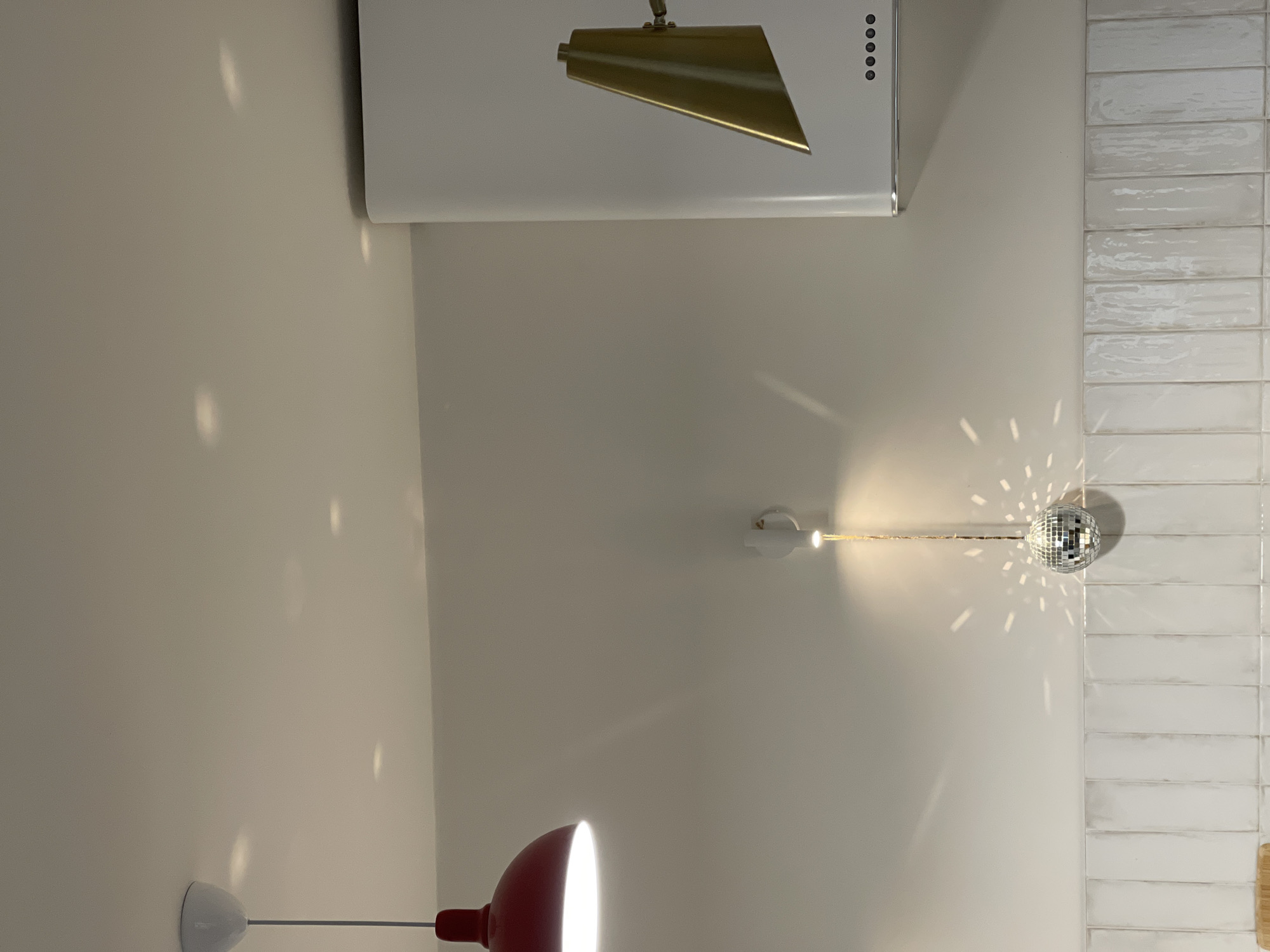 The image shows an interior space with white brick-like tiling to the right, a white exhaust hood with a gold accent above, and a red desk lamp at the bottom. A disco ball hangs near the tiling, reflecting light in a burst pattern. The walls are white. No people are visible. The image provides no indication of being in Izhevsk, Russia.
