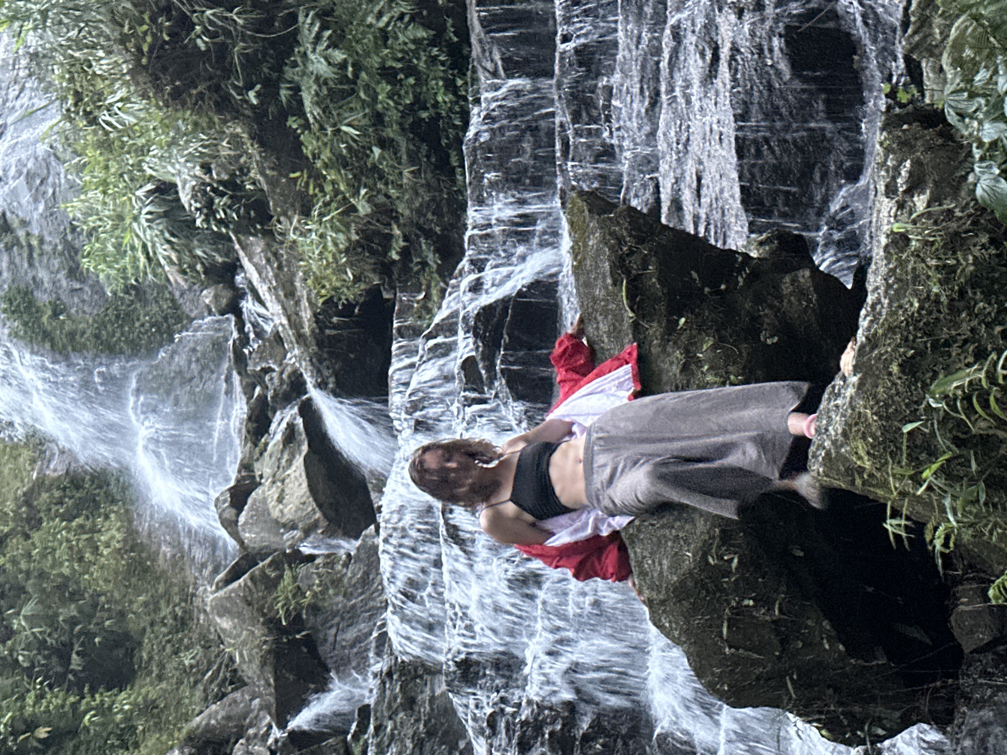 The image depicts a serene natural landscape in Thông Nguyên Commune, Vietnam, centered around a cascading waterfall and a person. A young woman, likely in her 20s or 30s, is seen relaxing on a large, dark rock to the right of the main waterfall. Her long, wavy brown hair partly obscures her face as she lies on her back or side. She wears a black tank top or bralette, grey loose-fitting pants, and a vibrant red and white garment draped around her, adding a pop of color against the natural tones.
The background features a powerful, multi-tiered waterfall, with white, frothy water flowing over dark, rugged rock formations. The rocks are prominently featured throughout the scene, appearing wet and covered in moss and various green plants. Lush green vegetation, including trees, bushes, and ferns, densely covers the surrounding cliffs and rocks, particularly on the left side and top of the frame, emphasizing the rich, natural environment.
The overall scene conveys a sense of calm and immersion in nature. The activity captured is one of relaxation or contemplation amidst the raw beauty of the waterfall. The lighting is soft and diffused, suggesting it is daytime, possibly overcast or shaded by the dense foliage, creating a tranquil atmosphere. There is no visible text present in the image.