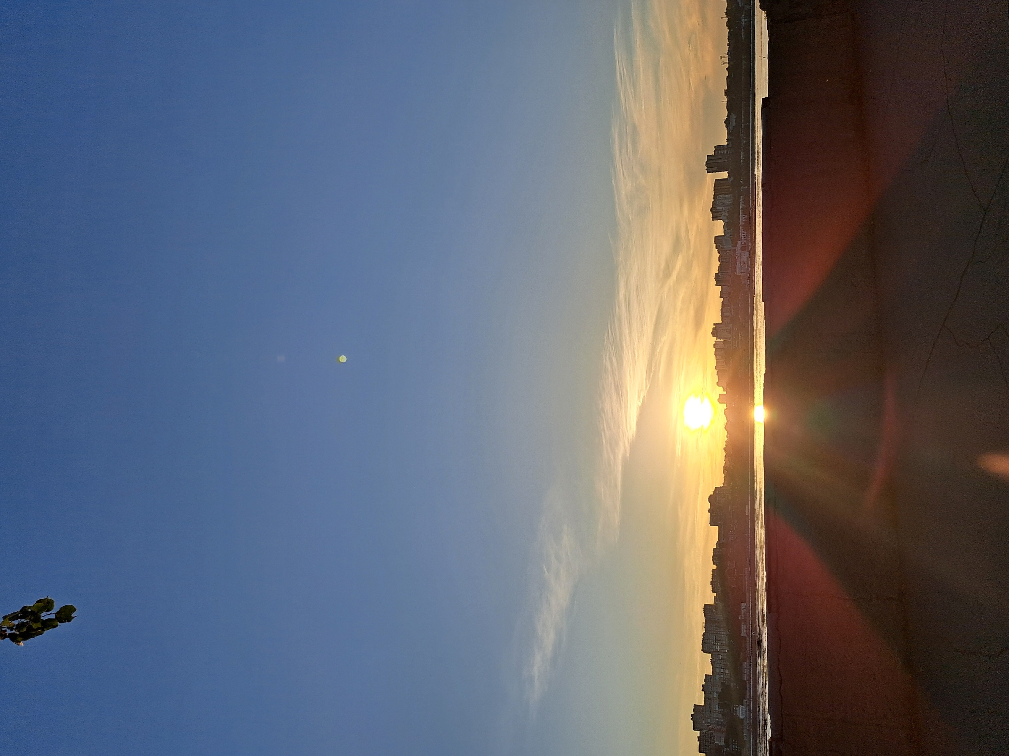 An outdoor, vertically oriented, eye-level shot captures a sunset over a city, likely Irkutsk, Russia, viewed from a waterfront promenade. The sky is a gradient of deep blue at the top, transitioning to pale blue and then to shades of yellow and orange near the horizon. Wispy clouds are scattered across the sky, particularly in the middle section. A single, small, bright object, possibly a distant airplane or drone, is visible in the upper left corner. Another faint, circular light artifact is present in the upper middle of the frame.
Below the sky, a cityscape is visible across a body of water, likely the Angara River. The buildings are silhouetted against the bright sunset, with many appearing to be multi-story residential blocks. The water reflects the warm colors of the sky. In the foreground, the top of a concrete or stone wall or embankment is visible, casting a long, dark shadow that stretches across the cracked asphalt of the promenade. The lighting suggests it is late afternoon or early evening. There are no discernible people or animals in the image. The overall atmosphere is serene and picturesque, highlighting the natural beauty of the sunset over an urban landscape.