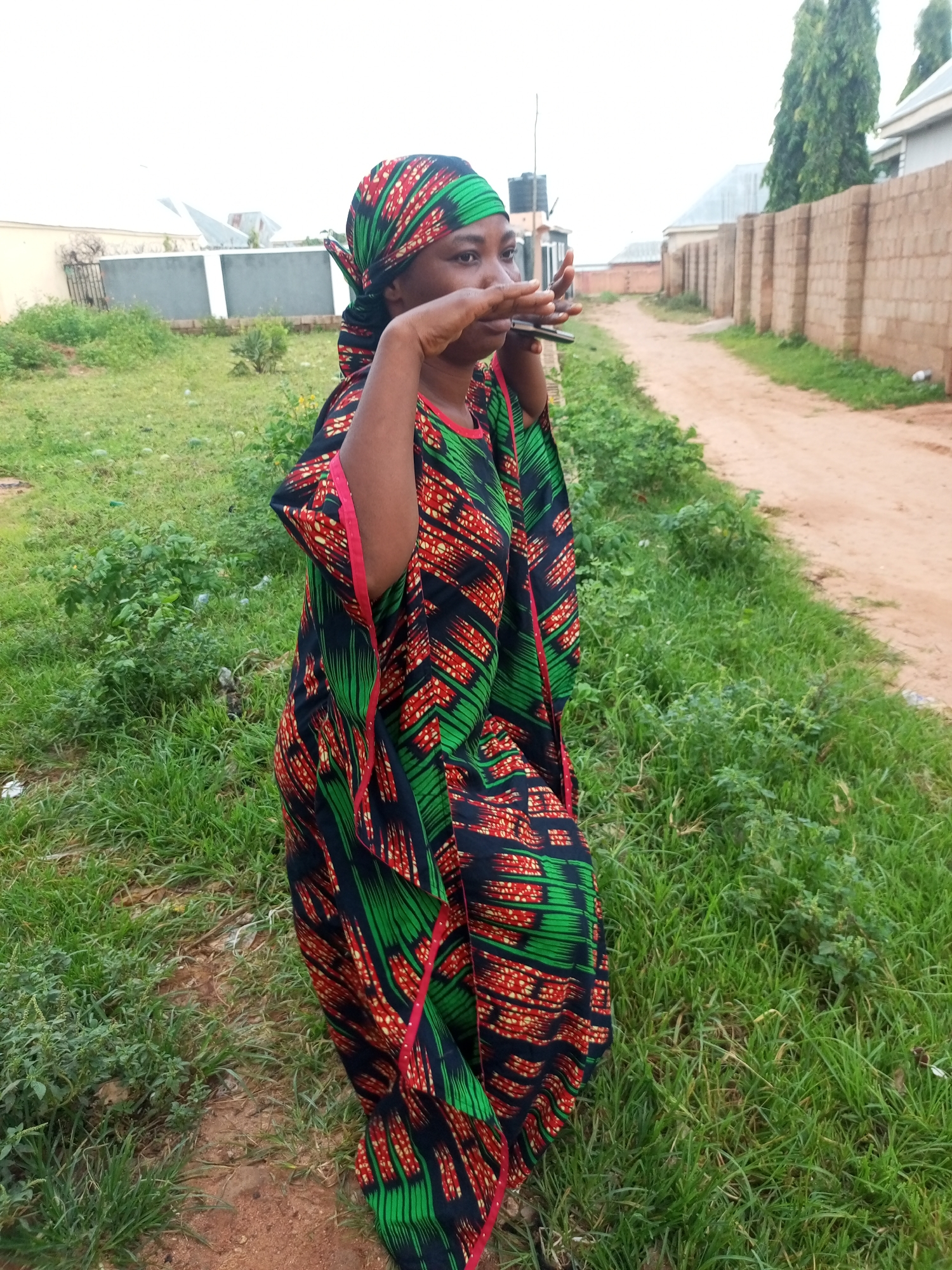 A medium-shot, outdoor, full-body, slightly right-facing photograph shows an adult female with dark skin wearing a vibrantly patterned green, red, and black headscarf and a matching flowing kaftan-style dress. Her right hand is raised, touching her mouth and chin as she looks off to the right of the frame, and her left hand is also raised. The woman is standing in an outdoor area with lush green grass and some patches of dirt. In the background, there is a dirt path, a low brick wall, and some buildings with neutral-colored walls and roofs. The sky is overcast.