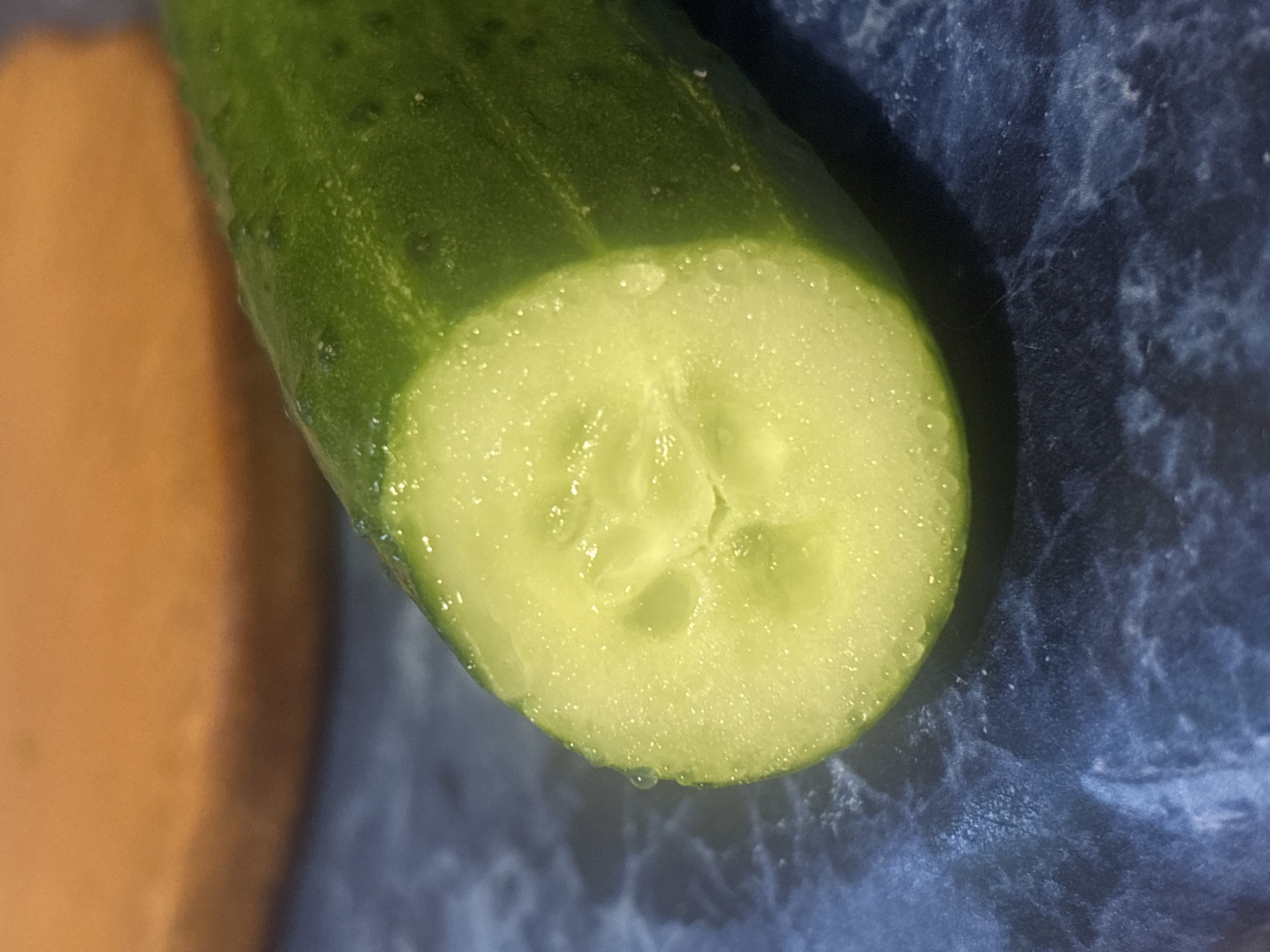 A close-up, high-angle shot shows a sliced cucumber on a dark blue, marbled surface. The cucumber is green with tiny bumps on its skin and water droplets on its surface. The sliced end reveals a pale green interior with seeds. In the background, out of focus, is an orange, circular object. This image does not contain any information about a specific location in Kremenchuk, Ukraine, nor does it depict any people or actions.