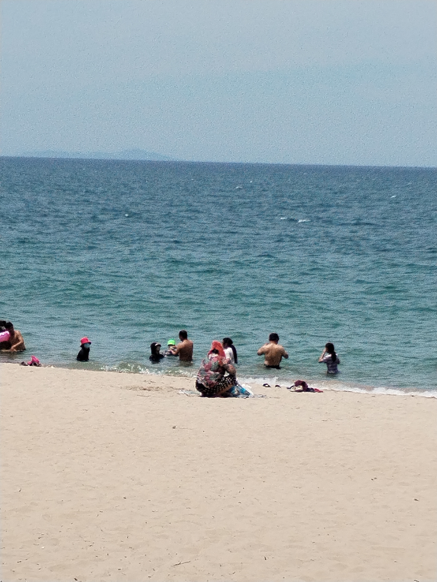 The image depicts a group of people at a beach in Nha Trang Ward, Vietnam. The scene includes a sandy shoreline in the foreground, with the ocean extending into the background. Several individuals are visible in the shallow water, some appearing to be swimming or wading. One person is sitting on the sand, facing away from the camera, and another is partially visible on the far left of the image, also in the water. The sky is a hazy blue, and a distant landmass is discernible on the horizon. The water is a moderate blue-green with some ripples.
