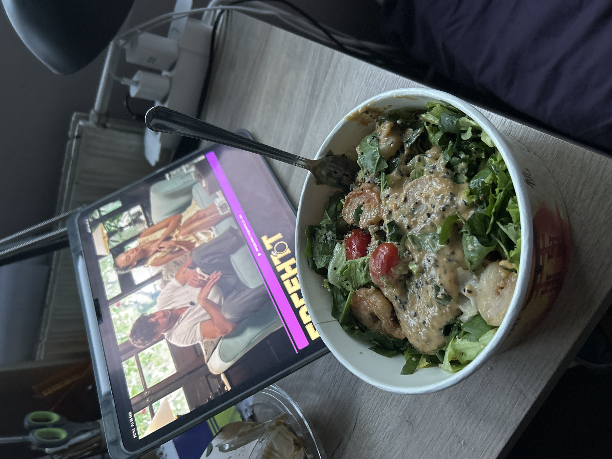 The scene depicts a tabletop setting in Sofia, Bulgaria. A white bowl filled with a salad consisting of lettuce, tomatoes, shrimp, and a beige-colored dressing with black specks is positioned on a light-colored wooden surface. A silver fork is partially submerged in the salad. To the left of the bowl, a tablet screen displays a scene of two people in what appears to be a movie or show. The name "Perchot" is visible on the screen. Adjacent to the tablet, there is a pair of scissors and a small clear container, possibly containing food. Above the scene, there is a complex of plugs and cords, suggesting a charging station. The background includes a dark purple cloth or fabric.
