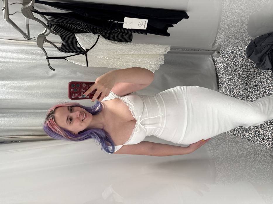 Inside a fitting room located in the City of Zagreb, Croatia, a female subject is depicted taking a mirror selfie. The subject, a young adult with long hair dyed purple and pink, wears a white, form-fitting, ribbed midi dress featuring a sweetheart neckline with lace trim and a front tie detail. She holds a smartphone with a patterned dark red case in her left hand, positioned to capture her reflection. To the subject's right, a metal clothing rail displays various garments, including a white textured lace top with black trim and several dark items, one bearing a visible "BERSHKA" tag. The room's walls are light-colored, possibly curtained, and the floor is covered with a dark, speckled carpet. A dark garment lies on the floor in the bottom right corner.