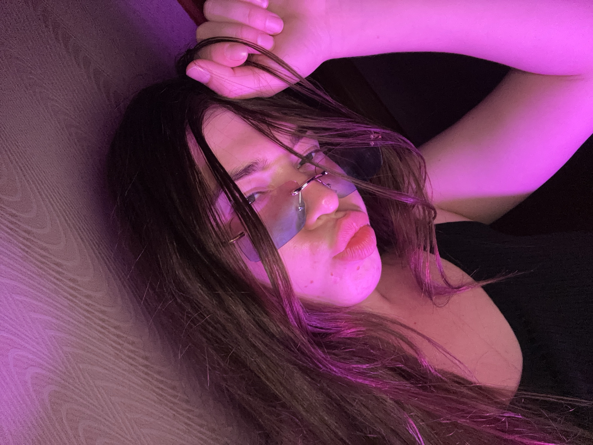 A young woman with long dark hair and light-colored skin is leaning her head against a textured surface, possibly a wall or ceiling. She is wearing a black top and rimless glasses with a metallic sheen. Her right hand is raised, and she is touching her hair near her temple. The scene is illuminated by pink light, creating a soft, diffused glow. Her lips are a bright pink color, and her eyes are visible through her glasses. The background appears to be a textured wall with wood grain patterns, bathed in varying shades of pink and purple light. The image is cropped closely, focusing on the woman's face and upper body. There is no visible indication of the location being in Мещёры, Russia.