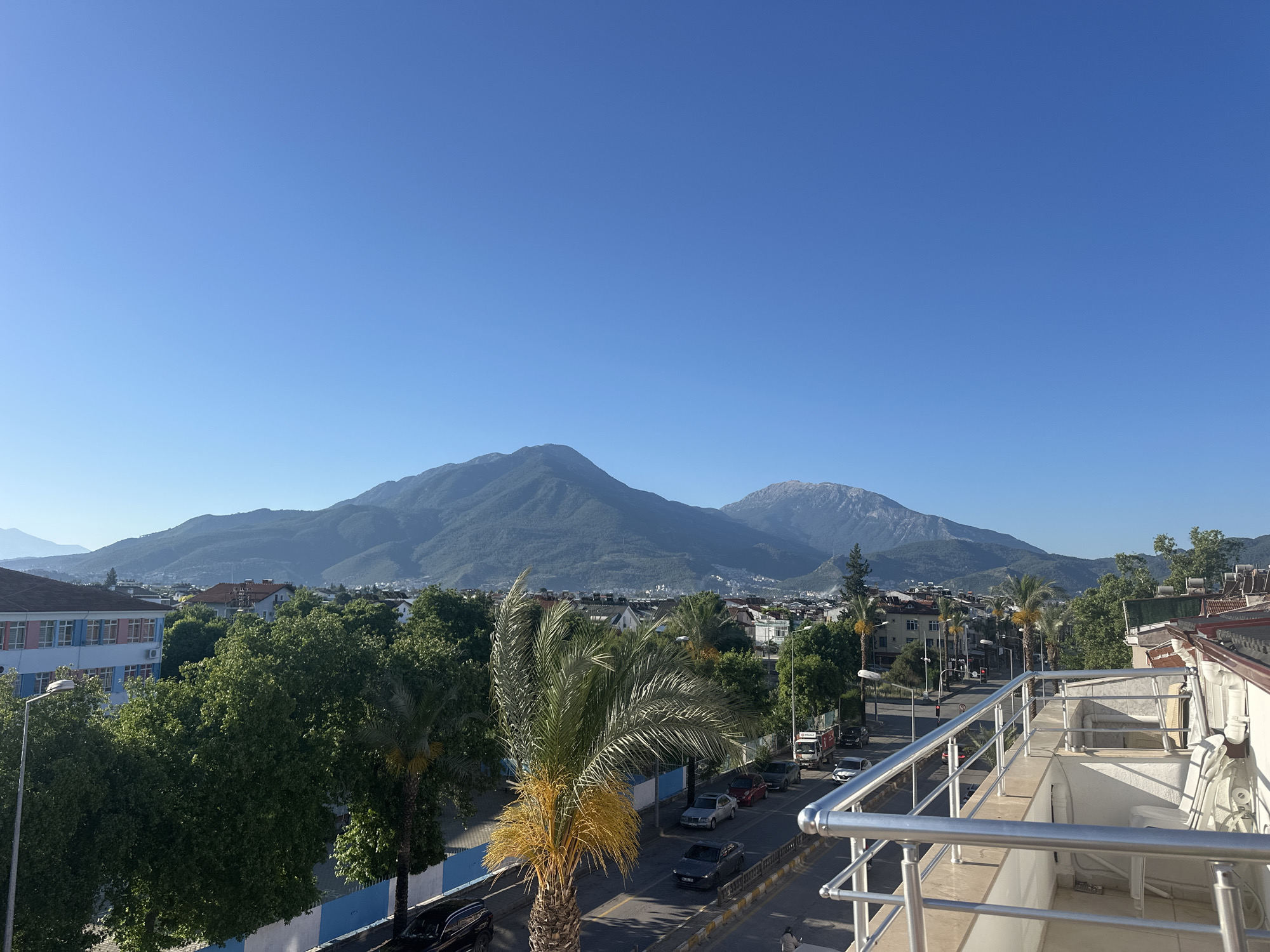 The image captures a cityscape in Fethiye, Turkey, featuring a clear blue sky and two mountains in the background. The foreground is dominated by a palm tree and a road with several parked cars. A building with a white and beige exterior, featuring a balcony with a metal railing, is situated on the right side of the image. On the left, there is a building with pink and light blue accents. Lush green trees and shrubs fill the space between the buildings and the road, adding to the urban landscape. The overall scene suggests a tranquil, sunny day in this Turkish city.
