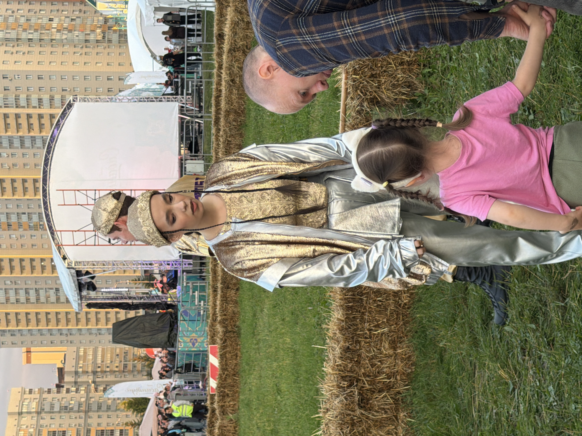 In an outdoor urban setting, two people in metallic gold and silver costumes stand near a stage in the background. A young girl wearing a pink t-shirt and olive green pants is in the foreground, facing away from the camera, holding hands with an adult male wearing a blue and brown plaid shirt. The adult male is looking down at the girl. The ground is covered in grass with hay bales placed strategically. In the background, tall buildings line the horizon, and a crowd of people is visible near the stage area.