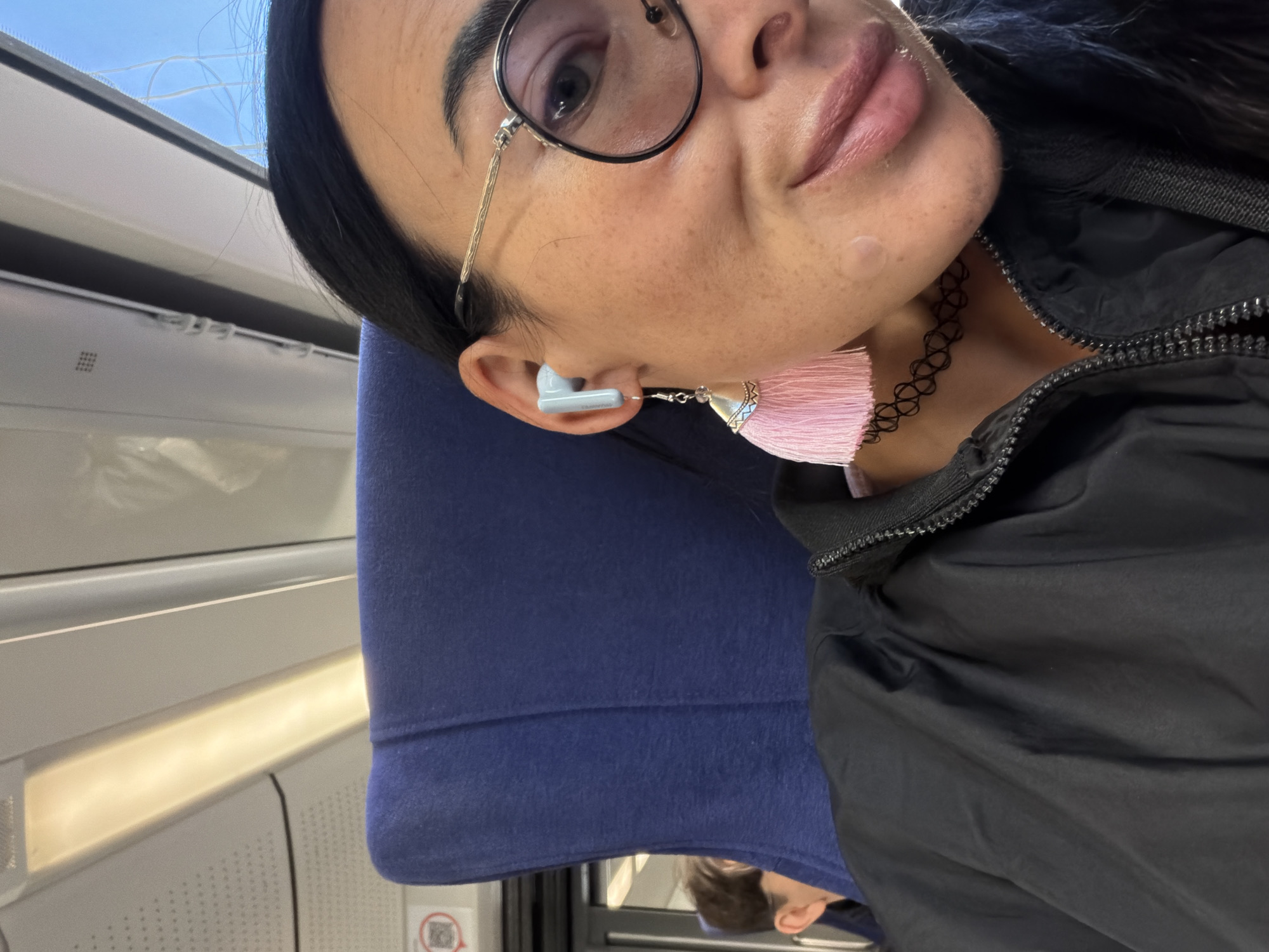 A woman with dark hair and glasses is seated in what appears to be a train or airplane, as indicated by the window and interior panels visible in the background. She's wearing a black jacket and has a choker necklace. The woman has a blue wireless earbud in her right ear with a decorative tassel dangling from it. The location within Saint Petersburg, Russia, is not discernible from the visual information provided.