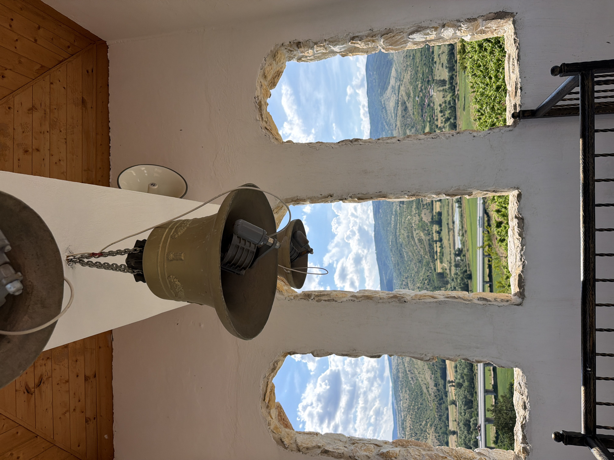 This image shows a view from inside a bell tower, looking out through three rough-hewn openings. The bell tower is white with a wooden ceiling. Two large bells hang from chains within the tower, the larger one in the foreground. Outside, a green, hilly landscape unfolds under a blue sky with white clouds. A highway bridge with vehicles can be seen in the middle ground. The scene is located in the vicinity of Kocherinovo, Bulgaria.