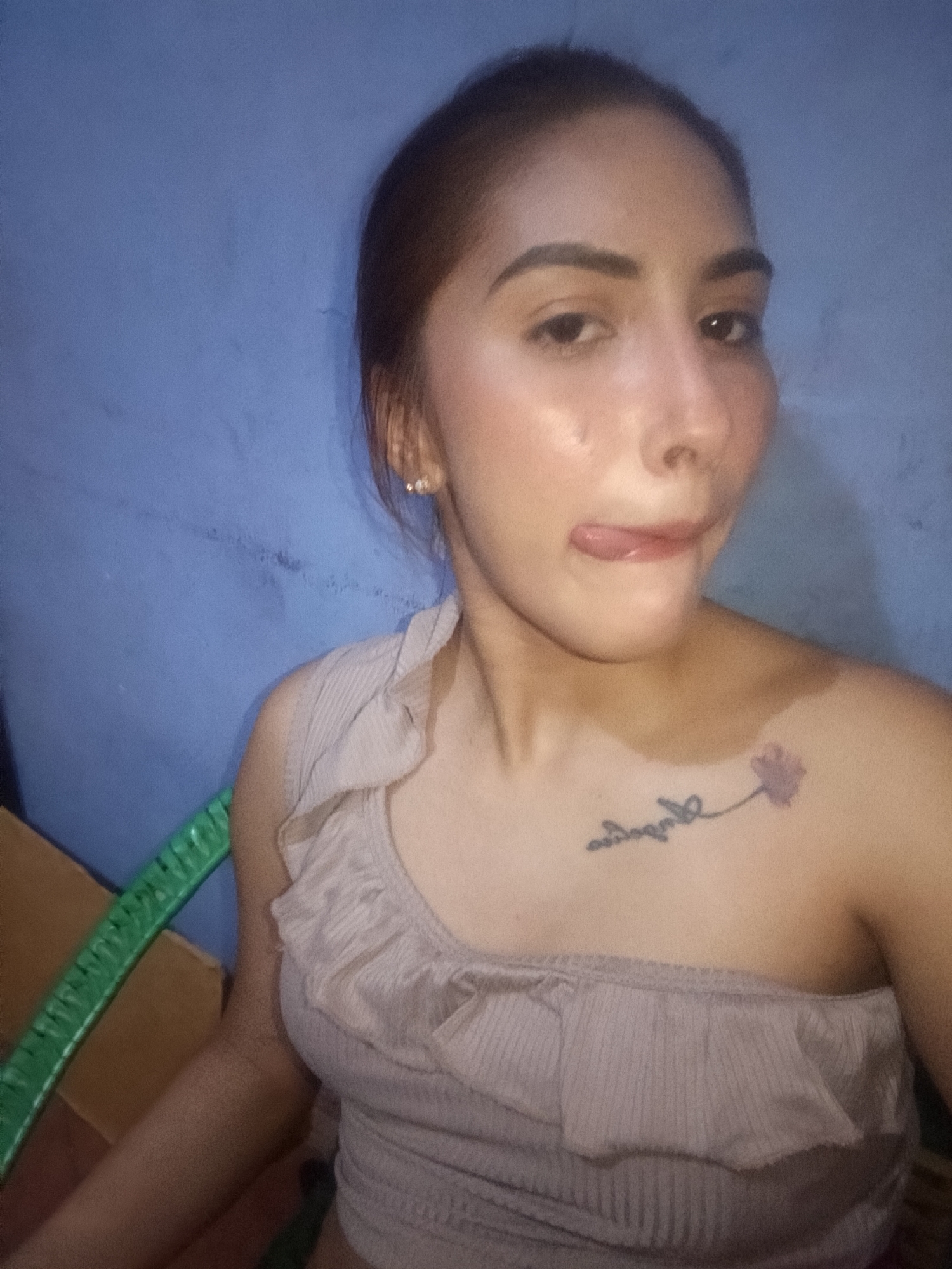 A woman with tan skin and brown hair pulled back is shown in a selfie-style shot. She is wearing a light brown one-shoulder top with ruffles and is sticking her tongue out slightly. She has a floral tattoo on her upper chest. The background is a blue wall. The photo was taken in Juan Griego, Venezuela. A green object is partially visible on the left side of the frame.