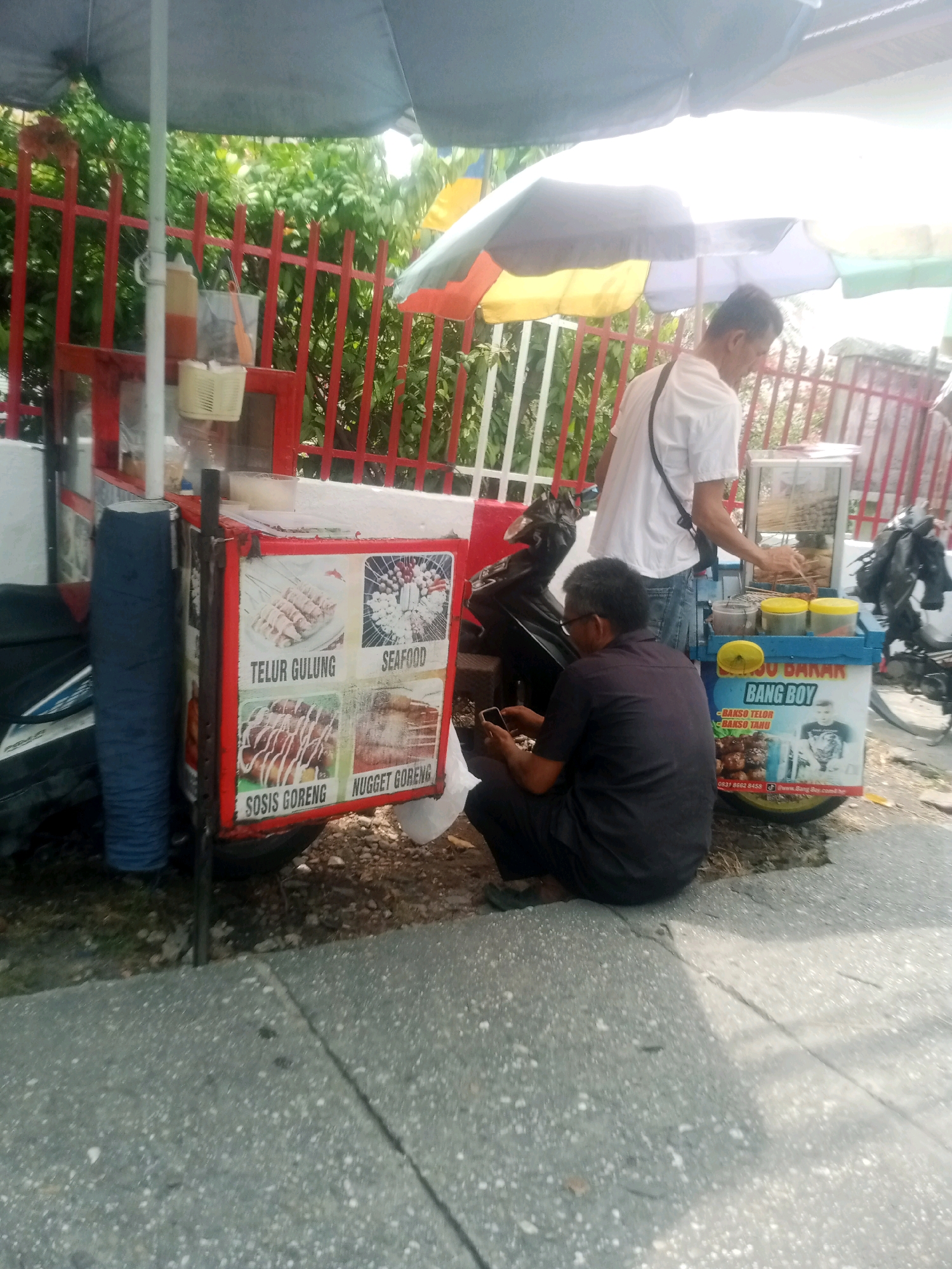The image depicts a street scene in Pekanbaru, Indonesia, featuring a food cart selling various fried snacks. Two men are present near the cart. One man, wearing a white t-shirt and jeans, is standing behind the cart, appearing to be preparing food. The other man, dressed in a dark shirt and pants, is squatting in front of the cart, looking at his mobile phone. The food cart displays images and text indicating menu items such as "TELUR GULUNG" (egg roll), "SEAFOOD", "SOSIS GORENG" (fried sausage), and "NUGGET GORENG" (fried nugget). There is also a label for "BANG BOY" with an image of a child. The environment includes a red metal fence, lush green foliage, and the ground is a mix of concrete and gravel. Umbrellas are set up over the food cart, suggesting it's an outdoor vendor. A motorcycle is parked next to the cart. The overall impression is that of a casual street food business operating in an urban setting.