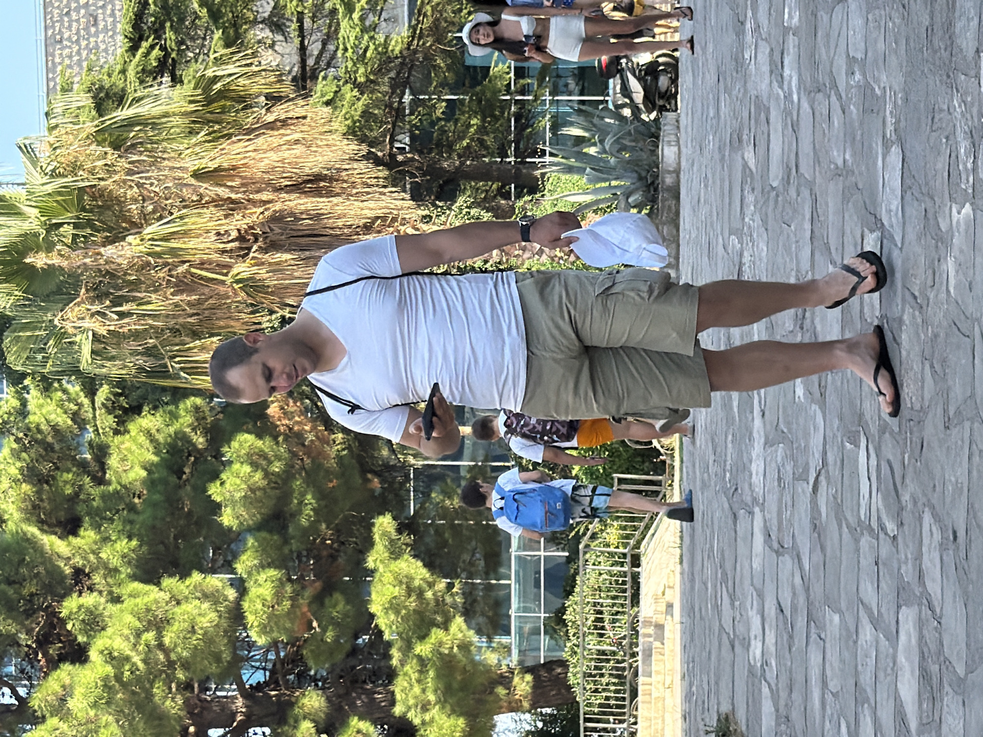 The image captures a sunny outdoor scene on a stone-paved pathway in Budva, Montenegro. In the foreground, a man is walking towards the viewer, looking down at his phone. He is wearing a white V-neck t-shirt, khaki cargo shorts, and black flip-flops. He carries a white folded item in his right hand. Behind him, two young boys are walking away. The boy on the left wears a blue backpack and patterned shorts, while the boy on the right wears an orange t-shirt and shorts. Further in the background, a woman in a white hat and white shorts is visible, along with other figures and a parked motorcycle. The environment is lush with trees, including a prominent palm tree, and appears to be a public area with some buildings in the distance.