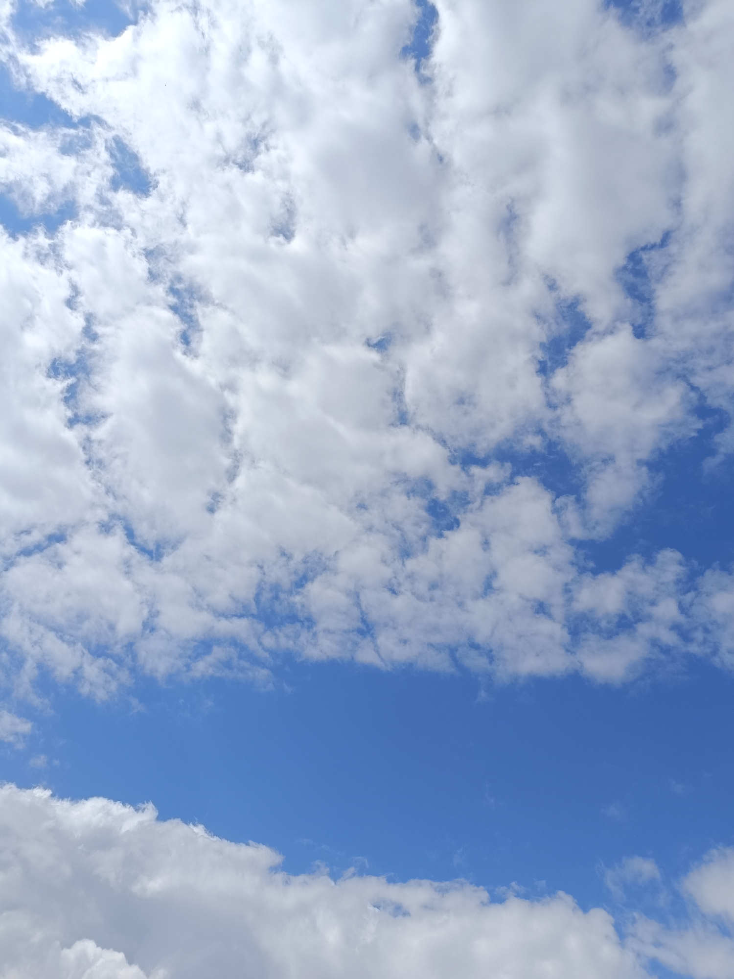The image shows a daytime sky with a mix of blue sky and white, puffy clouds. The clouds are scattered throughout the frame, with denser clusters in the upper portion and bottom edge. A clearer patch of blue sky stretches across the middle of the image. No people, subjects, or objects are visible other than the sky and clouds. There are no actions or interactions depicted. The environment is simply the sky, observed during daylight hours. No specific location in Vasil Drumev, Bulgaria, can be determined from the image.