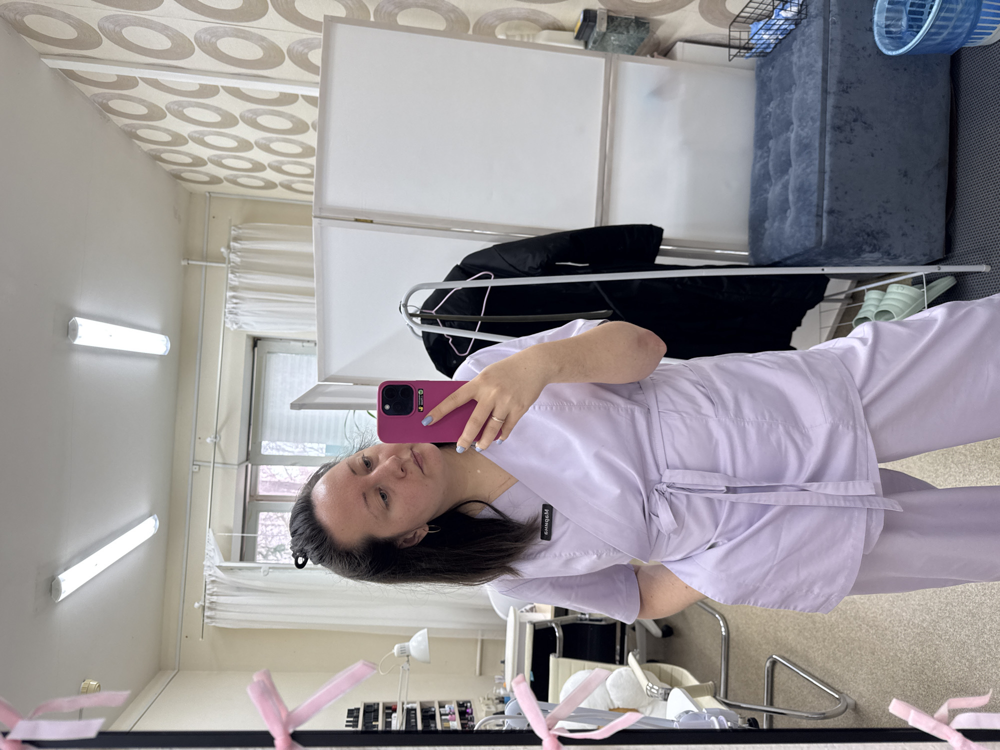 A woman is captured taking a mirror selfie in an indoor setting, likely a professional environment such as a clinic or salon. She has dark hair and is wearing a light purple medical scrub top with matching pants. In her left hand, she holds a pink smartphone encased in a dark cover, showcasing light blue nail polish on her fingers and a simple silver ring. A name tag with mirrored and inverted text, possibly "МdФНН6" or a similar Cyrillic word, is pinned to her scrub top.
The room's reflection in the mirror reveals various details. Behind the woman, a white, multi-panel room divider or screen is visible. To her right, a dark jacket hangs on a metal rail, and further right, a dark blue upholstered storage ottoman or bench sits on a light-colored carpeted floor. A pair of light green slides or slippers are placed near the ottoman.
To the left of the woman's reflection, a window with white horizontal blinds shows blurred outdoor scenery, suggesting foliage. A horizontal white fluorescent light fixture is mounted on the wall. The ceiling features a distinctive wallpaper pattern of light brown or gray concentric circles.
In the immediate foreground, closer to the mirror's bottom edge, parts of a white treatment chair or bed with metal legs are visible. Below this, shelves or a cart display numerous small bottles and containers, indicative of cosmetic or medical supplies. Several pink ribbons are tied to the top edge of the mirror frame. The lighting is bright, and the woman maintains a neutral expression.