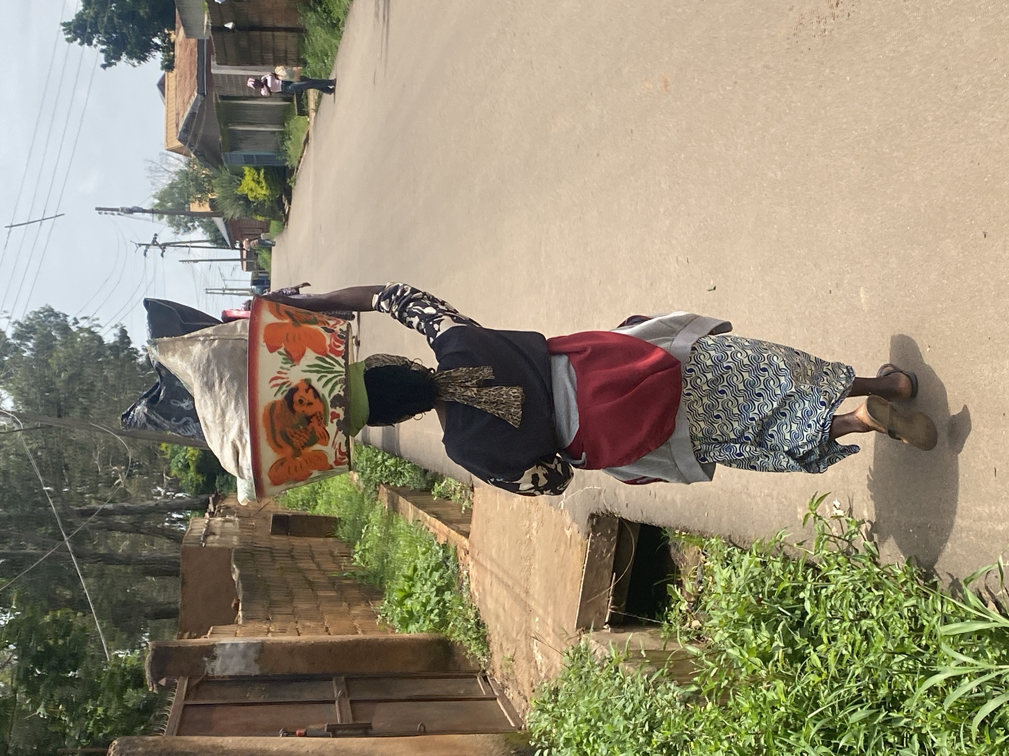 In Gura Top, Nigeria, a woman walks down a paved street carrying a large, decorated bowl filled with items on her head. The bowl is white with orange and red fish and floral designs. She wears a dark long-sleeved shirt with a white and black pattern on the sleeves, a red garment tied around her waist, and a blue and white patterned skirt. She also has a light-colored scarf tied around her head and neck. The street is lined with buildings on the right and a brick wall with a metal gate and overgrown vegetation on the left. The sky is overcast, and there are trees and utility poles visible in the background. Other people are walking in the distance on the street.