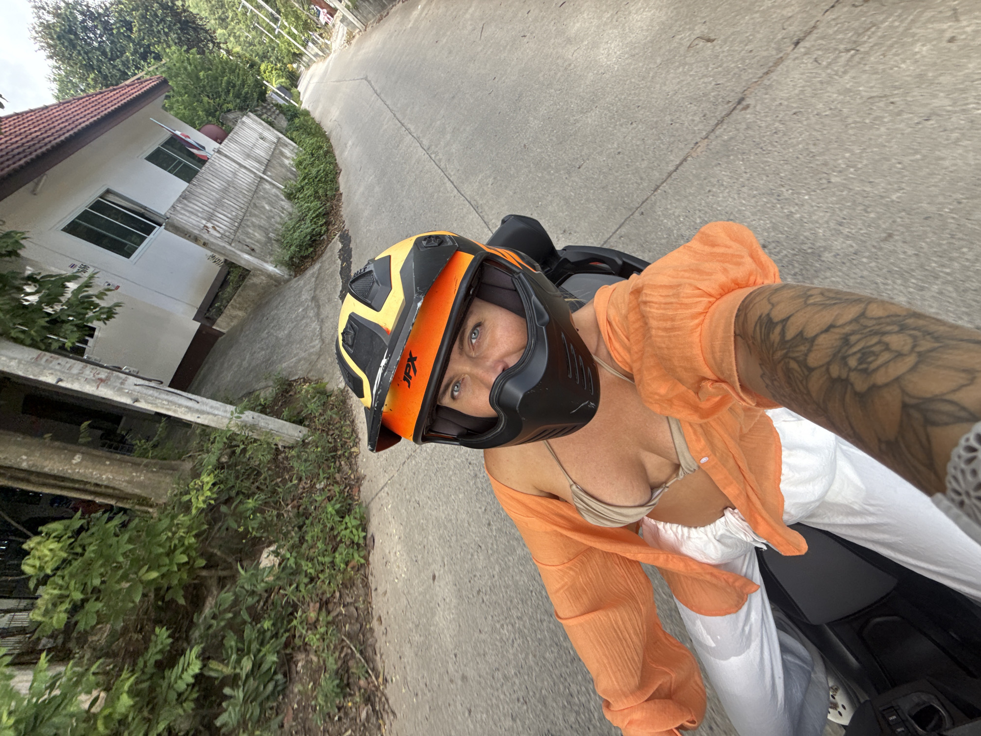 A woman wearing an orange cover-up over a bikini top and white pants rides on a motorcycle, facing the camera. She has a black and yellow helmet on and is looking at the camera with blue eyes. She is on a paved road in Ko Samui District, Thailand, with a white building and greenery on the side of the road. The image is taken from the perspective of the person driving the motorcycle.