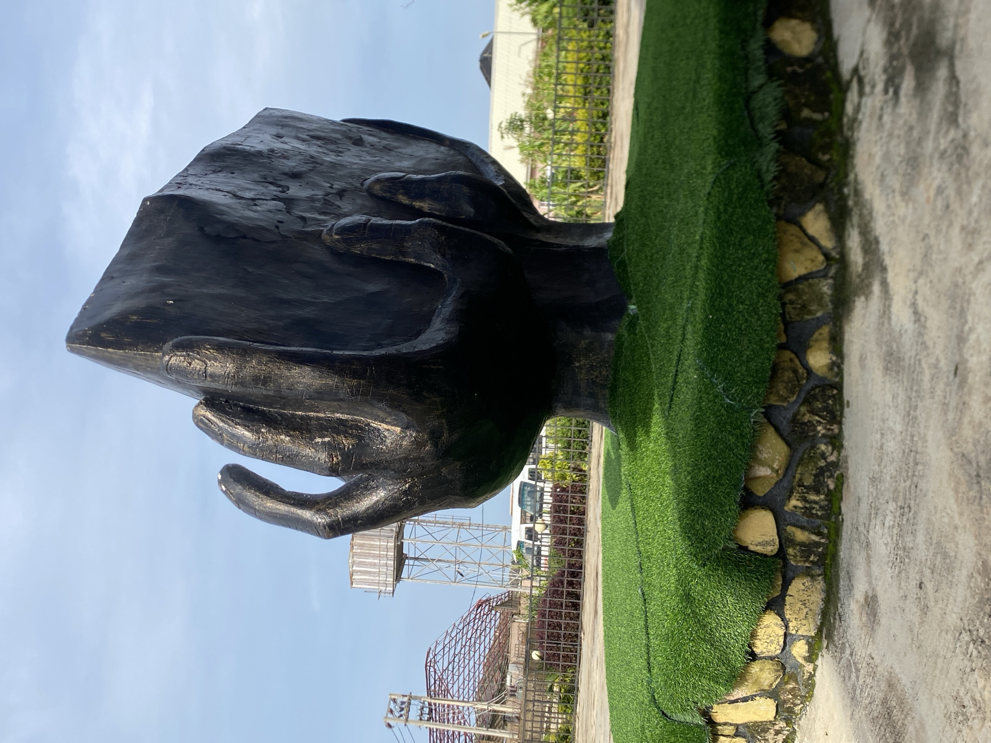 This is a medium shot, eye-level, outdoor photograph of a large, dark-colored sculpture in the city of Jos, Nigeria. The sculpture depicts two hands holding a rectangular object, possibly a book or tablet, and is situated on a circular raised platform covered with artificial grass. The base of the platform is constructed from uneven, yellow-toned stones. In the background, a tall water tower stands against a bright blue sky with scattered clouds. A metal-framed structure, possibly under construction or a skeletal framework, is visible to the left, along with a fence and some greenery. Vehicles, including a white bus, are parked behind the fence. The ground in the foreground is a light-colored concrete pathway.